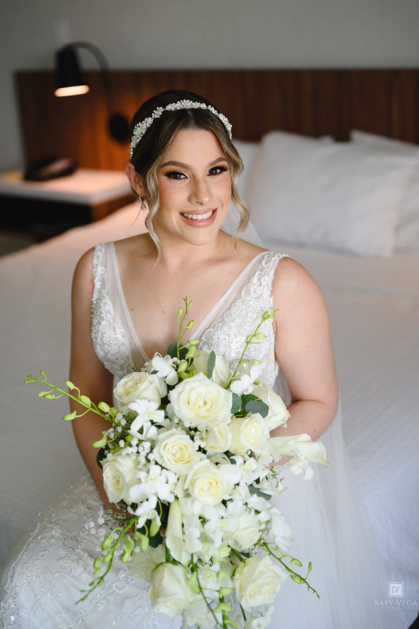 Bride in white lace gown holding cascading bouquet of white roses, smiling in hotel room.