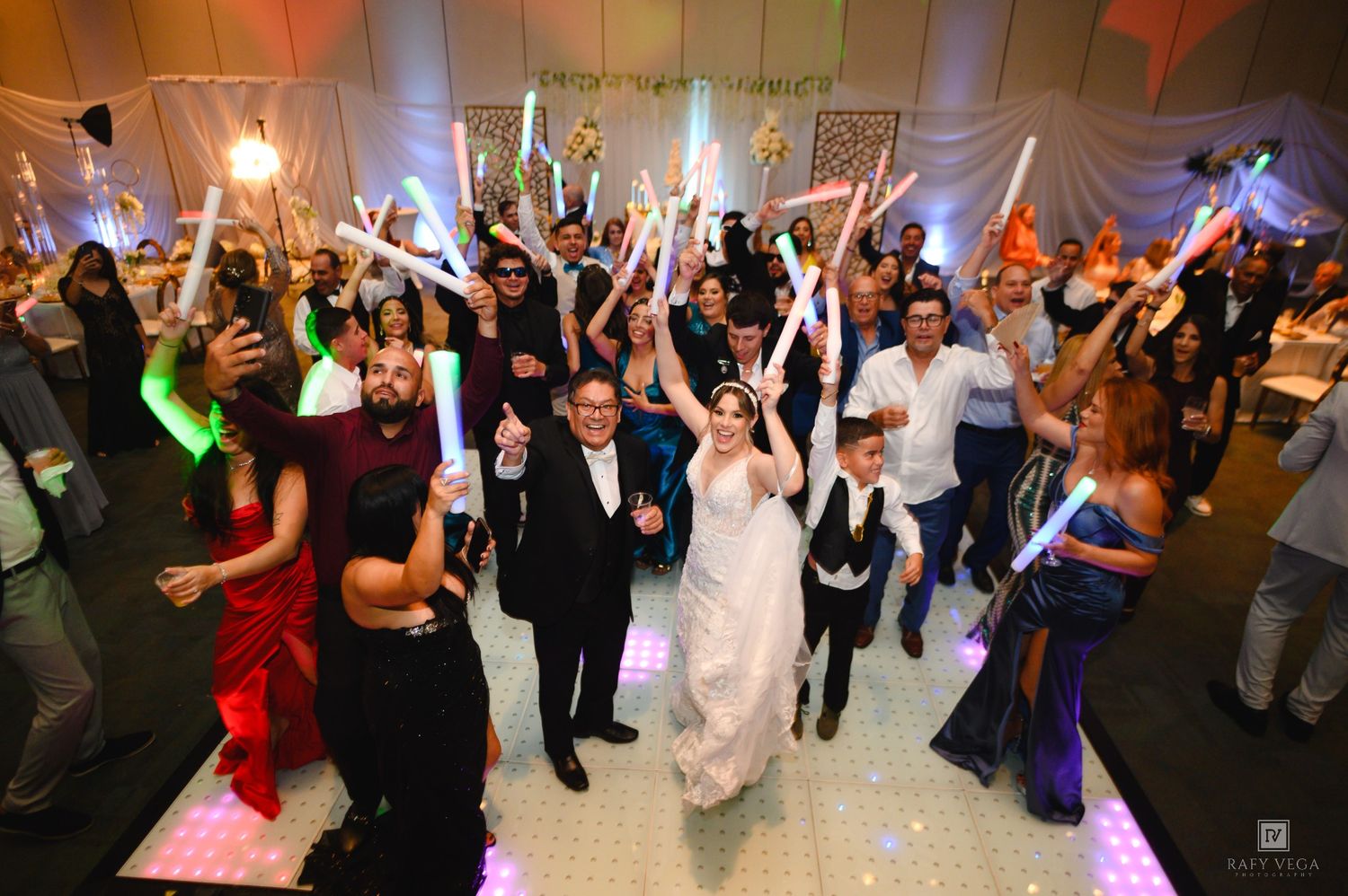 Joyful wedding reception with bride and groom surrounded by guests waving colorful glow sticks on a white dance floor.