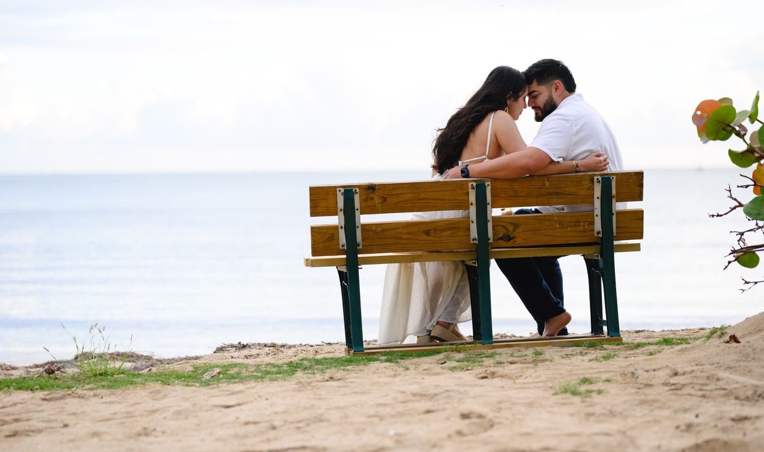 Couple sitting close together on a wooden bench by the ocean shore on a sandy beach.