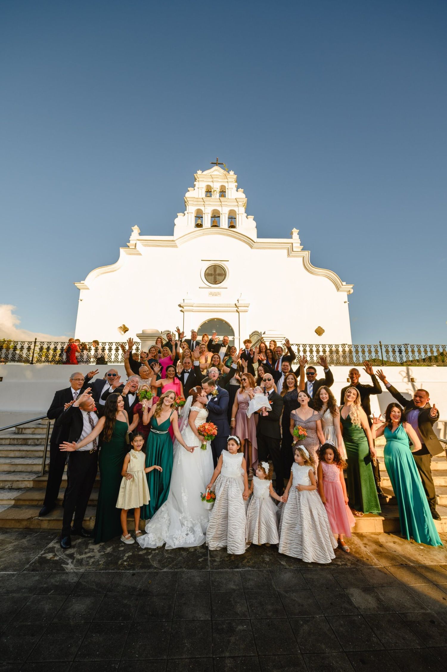 Wedding party poses joyfully in front of a white colonial church with bell tower on a sunny day.