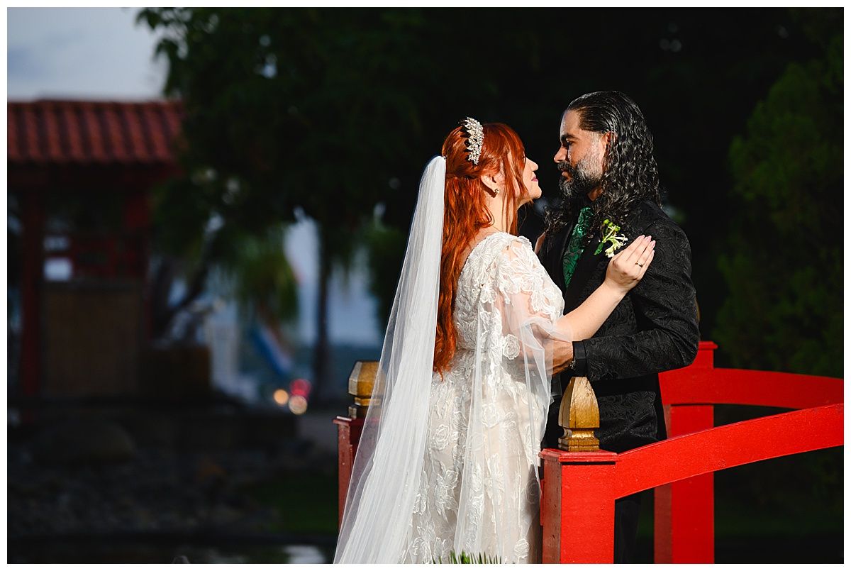 Bride and groom share a romantic moment at an outdoor wedding ceremony on a red platform surrounded by lush greenery.