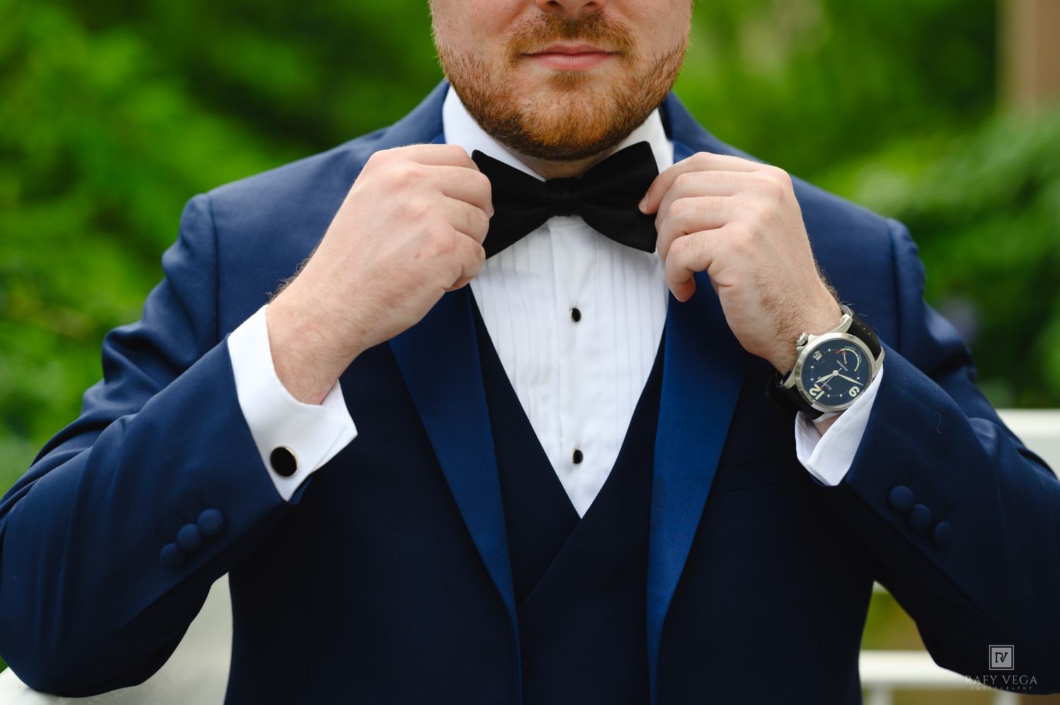 A groom in a navy blue tuxedo adjusts his black bow tie, wearing a watch and cufflinks outdoors.