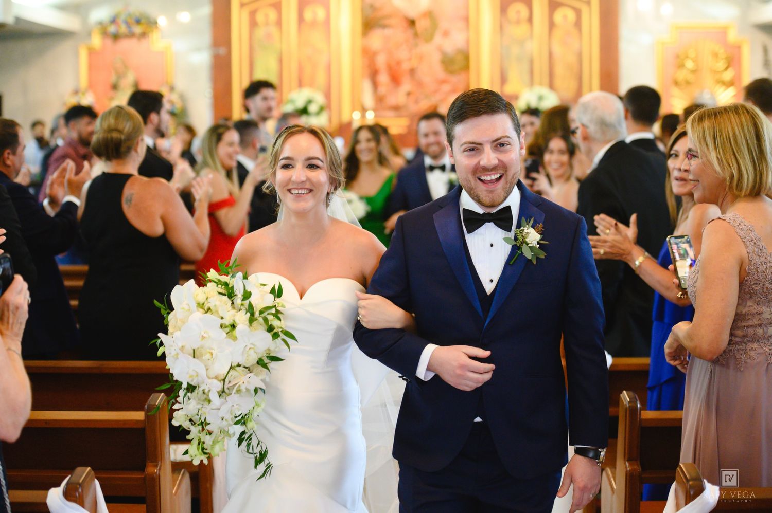 Bride and groom walk down church aisle smiling, bride holding white floral bouquet, groom in navy tuxedo.