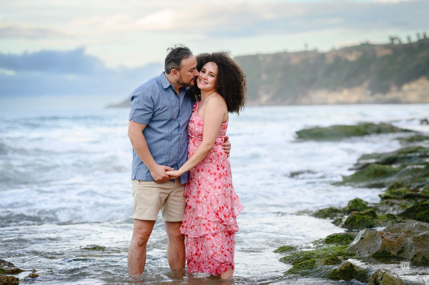 Couple embracing romantically on rocky beach shore with ocean waves, woman in pink floral dress, man in blue shirt.