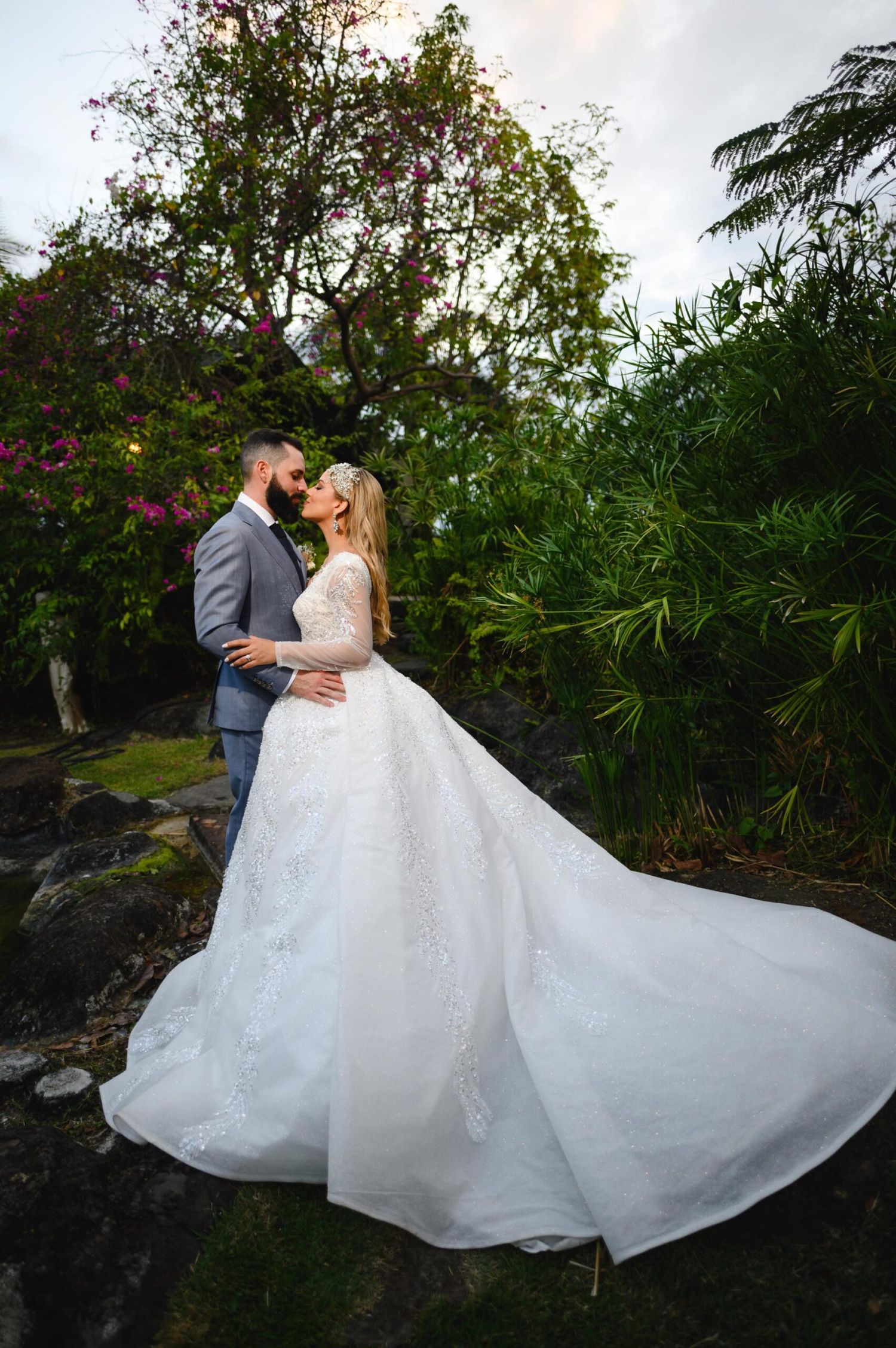 Bride in white lace ball gown kissing groom in gray suit outdoors surrounded by lush tropical greenery and pink flowers.