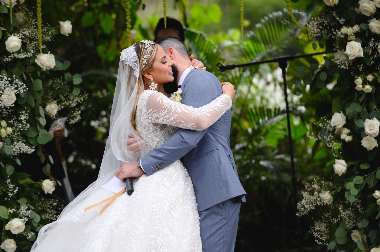 Bride and groom share first kiss under white floral wedding arch surrounded by lush greenery.