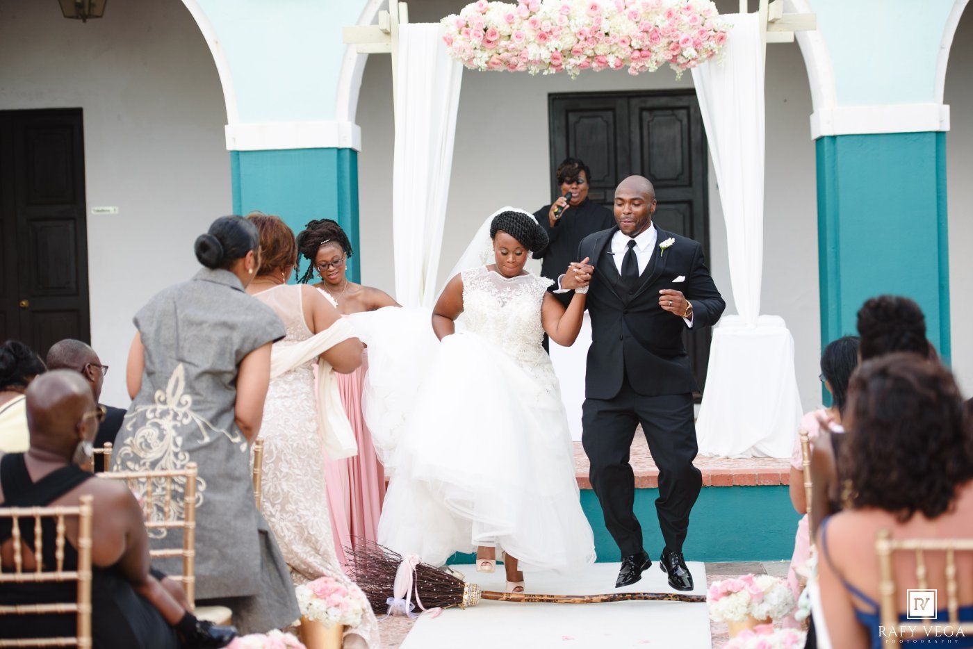 Bride and groom celebrate at outdoor wedding ceremony with floral arch, guests seated on gold chairs.