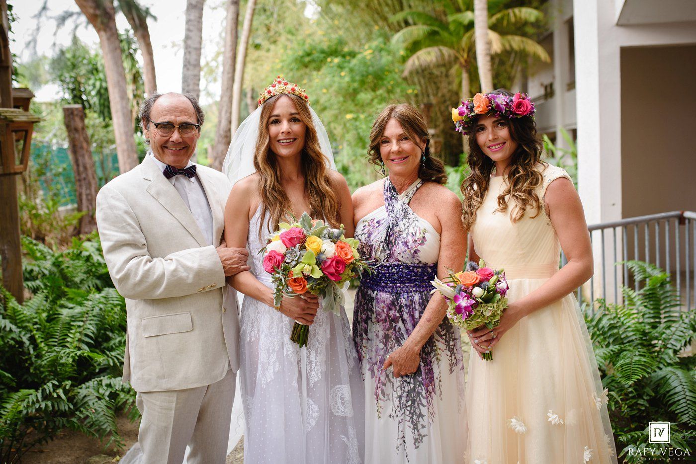 Family portrait with bride in white gown, floral crown, colorful bouquets, outdoors in lush garden setting.