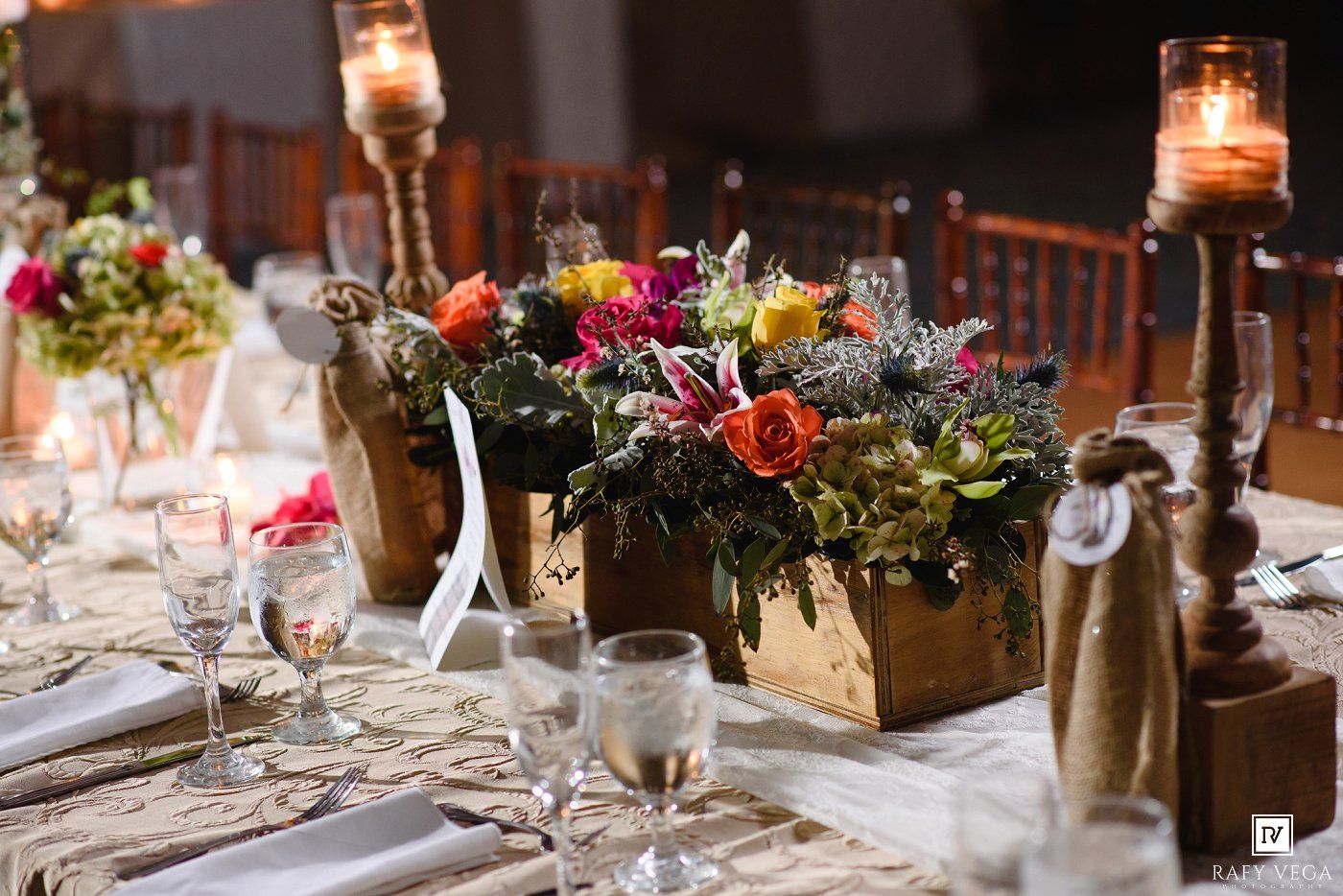 Rustic wedding reception table with colorful floral centerpieces in wooden boxes, candles, and elegant place settings.