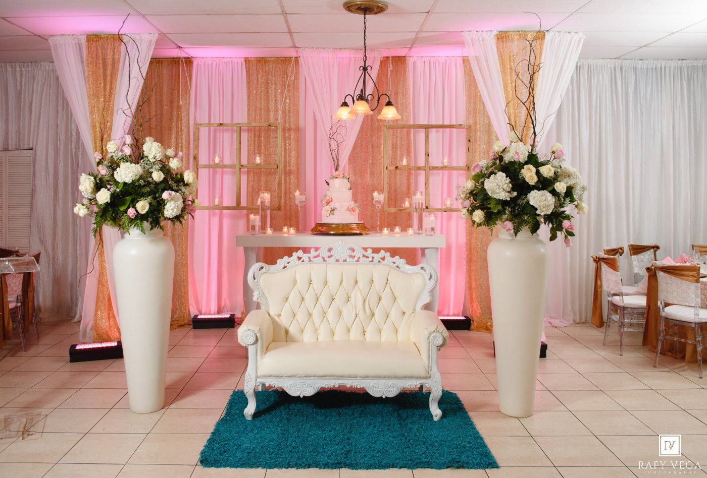 Wedding reception venue with white tufted loveseat, tall floral arrangements, cake table, and pink draped backdrop.