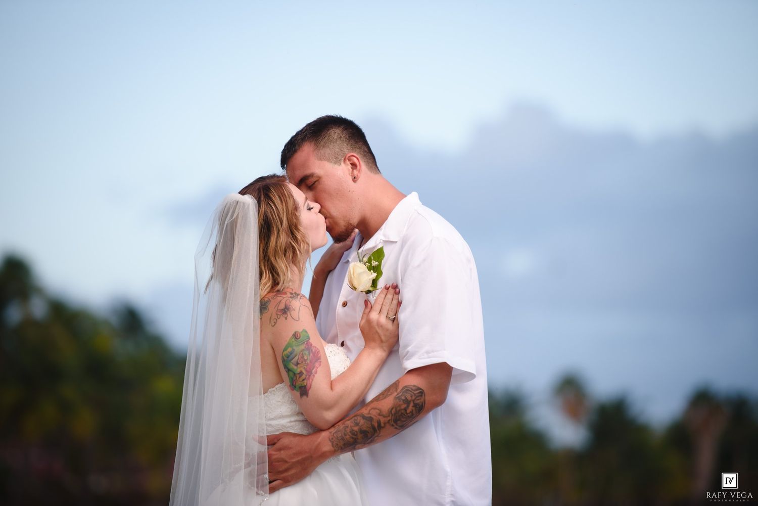 Bride and groom sharing a romantic moment at a tropical beach wedding, both wearing white with visible tattoos.