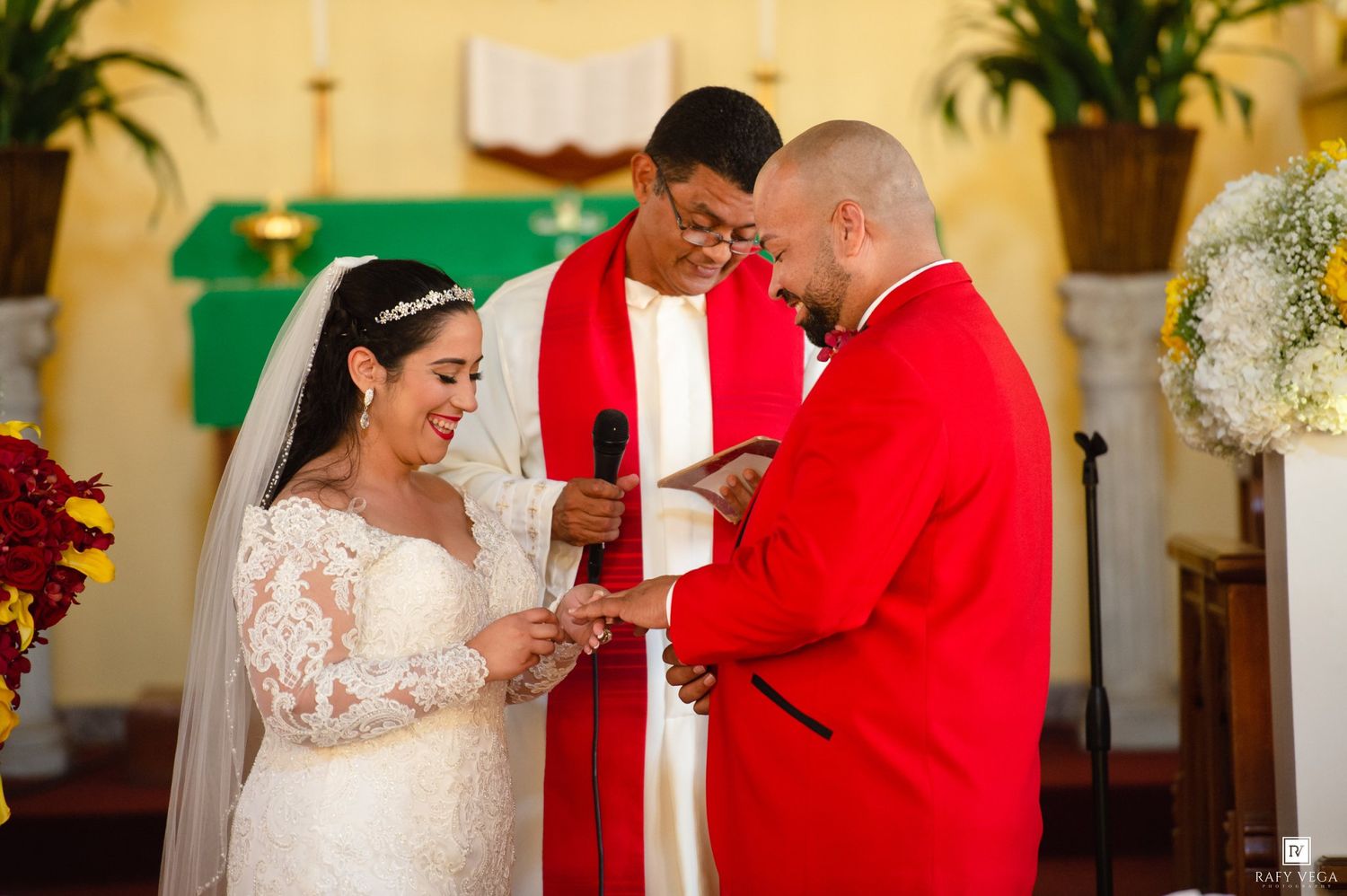Bride and groom exchange rings at church altar, officiant in red stole holds microphone during wedding ceremony.