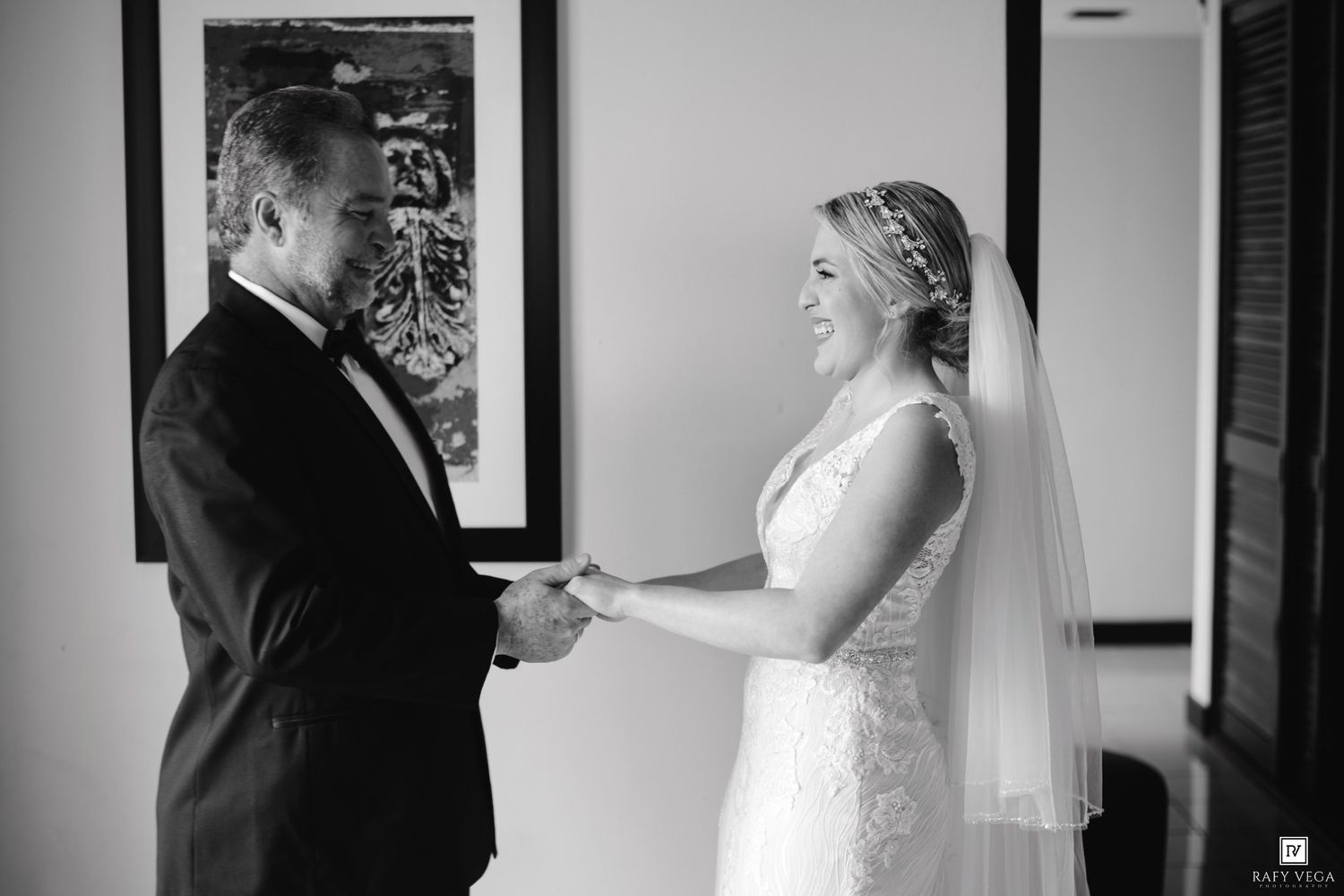 Black and white photo of a bride in lace gown and veil holding hands with a man in a suit indoors.