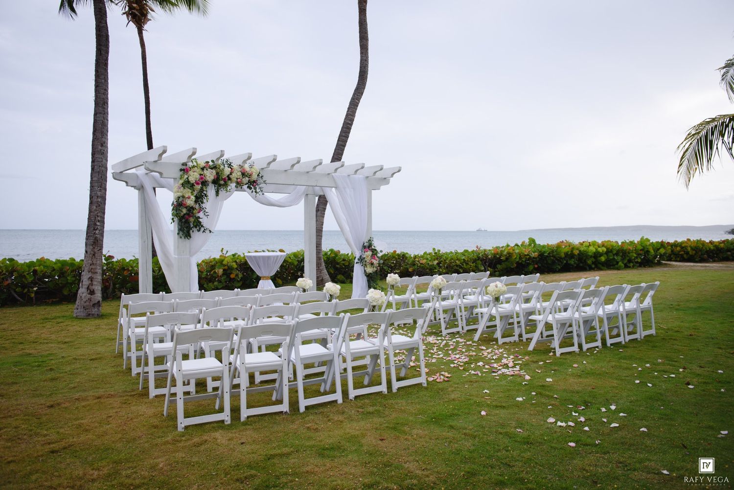 Elegant outdoor wedding ceremony setup with white chairs and floral arch overlooking the ocean on a tropical lawn.