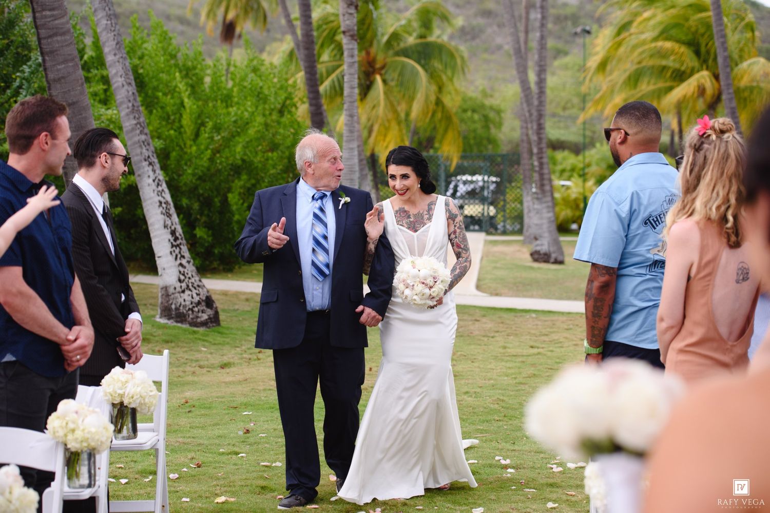 Radiant bride in white lace gown walks down outdoor aisle with older gentleman amid tropical palm trees.