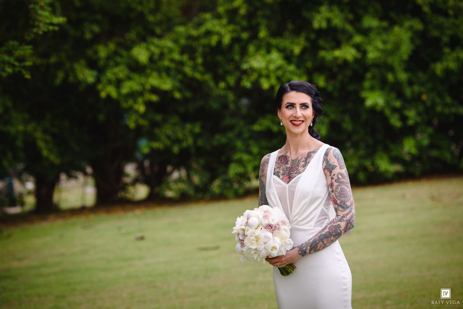 Tattooed bride in white wedding dress holding white floral bouquet smiling outdoors on green lawn.