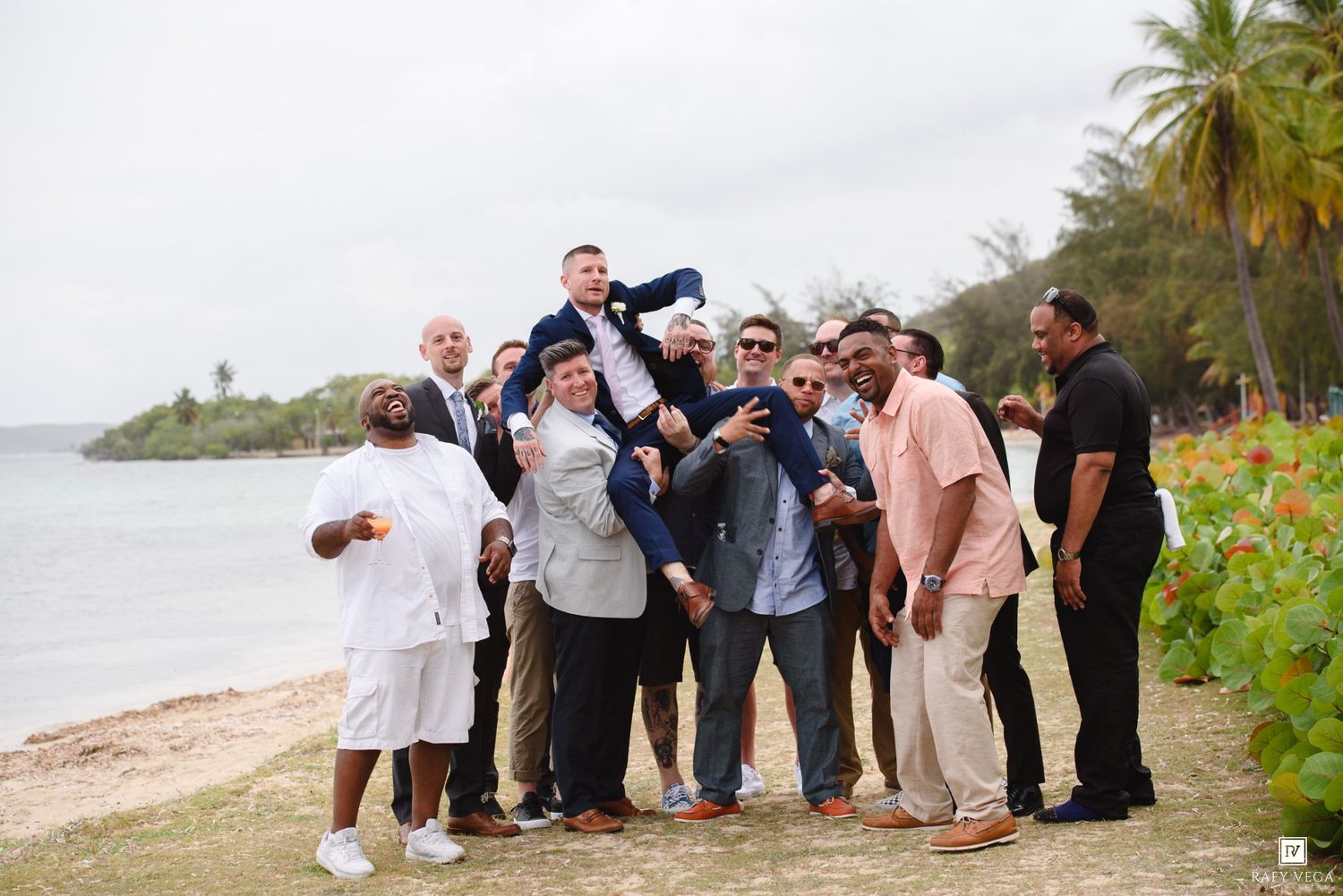 Groomsmen lifting groom on tropical beach during wedding celebration with palm trees and ocean in background.