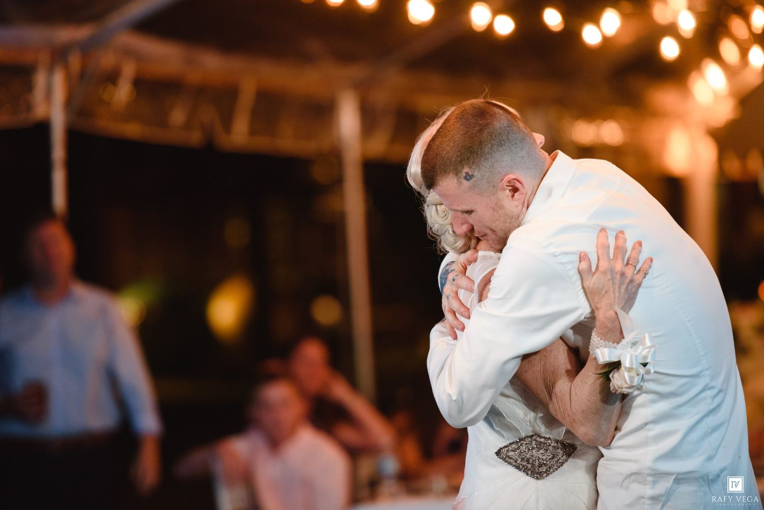 Groom embracing a guest in an emotional hug during a warm outdoor wedding reception under string lights.