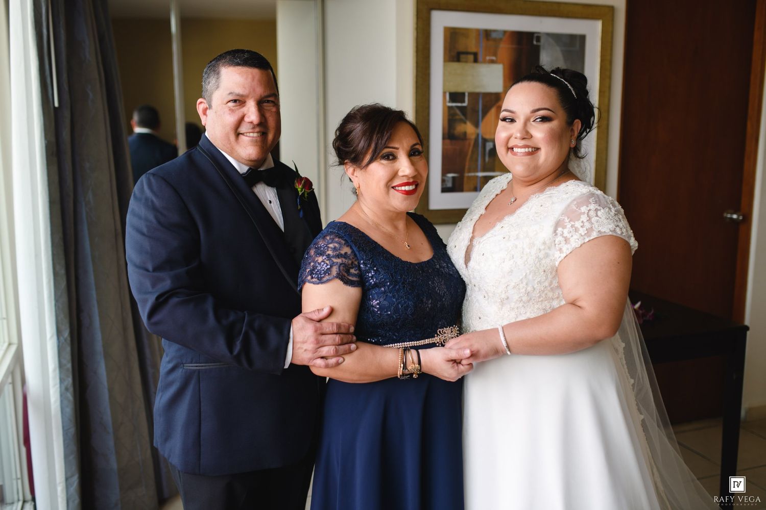 Bride in white lace gown poses with man in navy tuxedo and woman in blue dress at wedding venue.