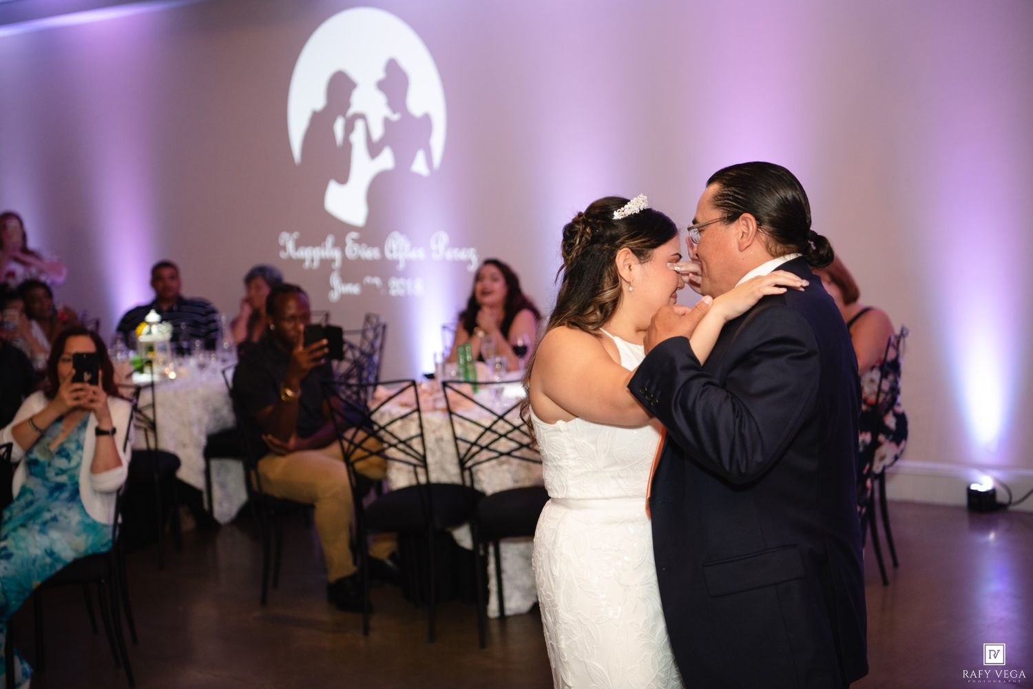 Bride and groom share their first dance at a wedding reception with purple lighting and guests watching.