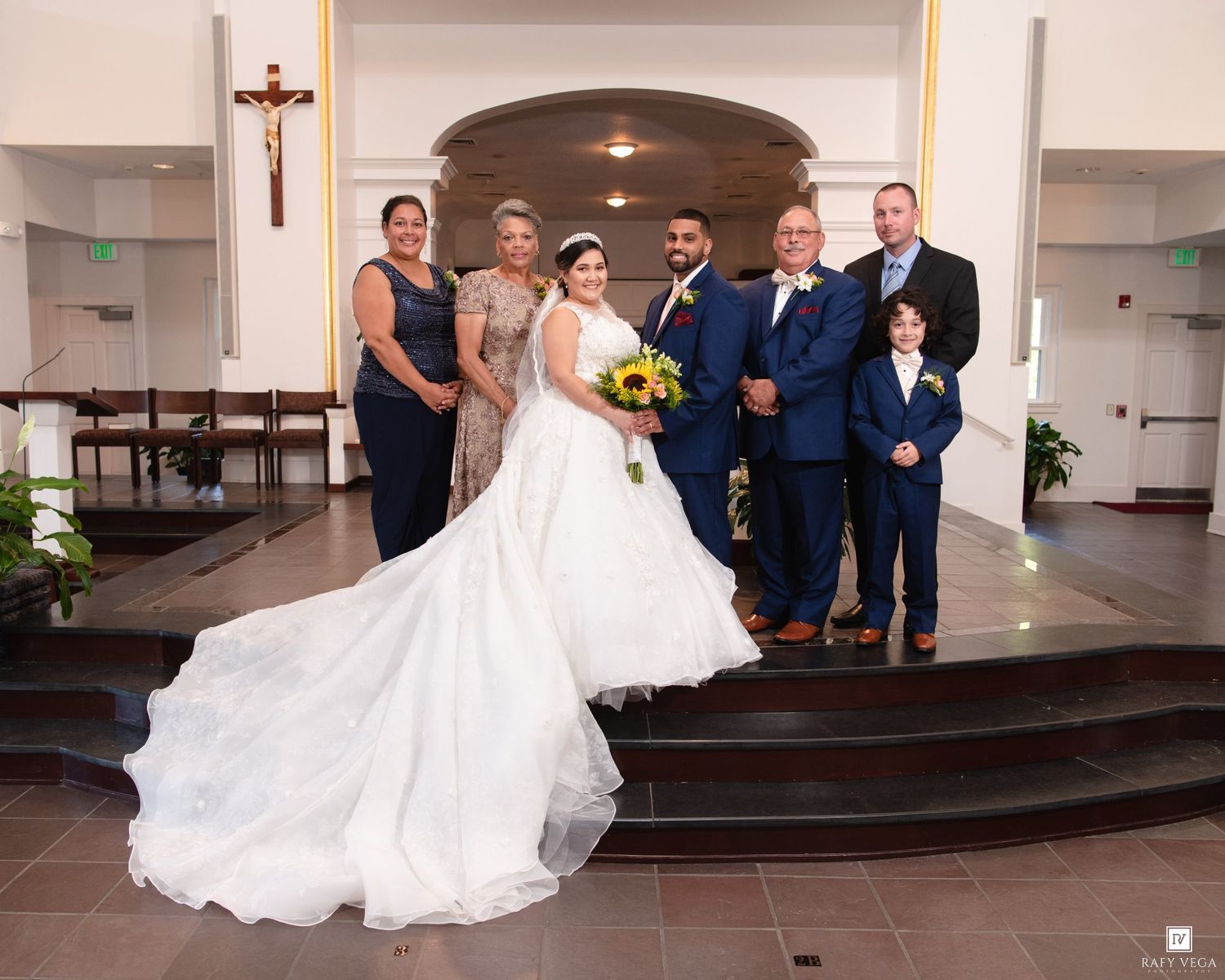 Wedding party poses at church altar with bride in ball gown holding sunflowers after Catholic ceremony.