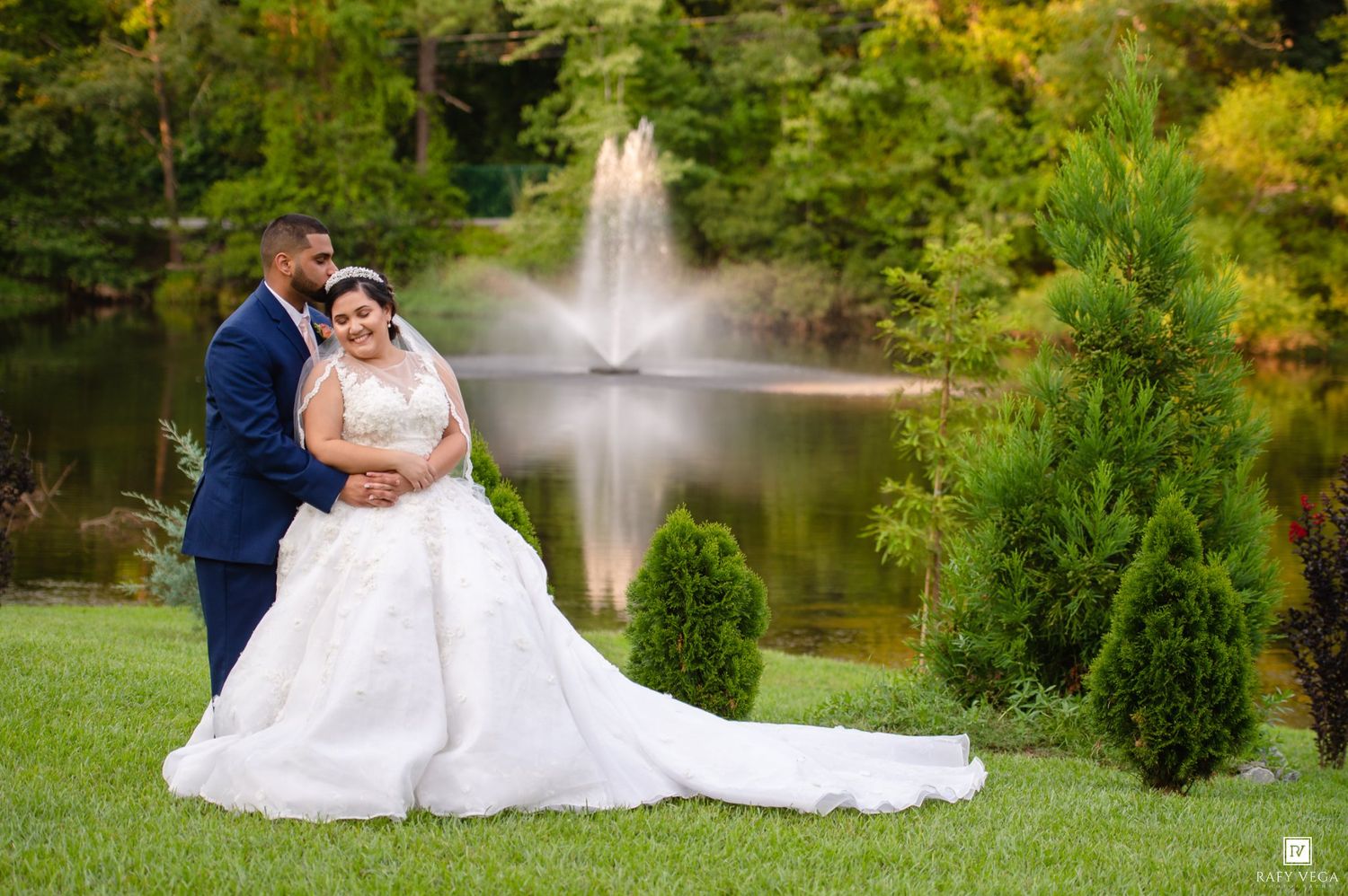Bride and groom posing by a serene pond with a fountain and lush greenery on their wedding day.