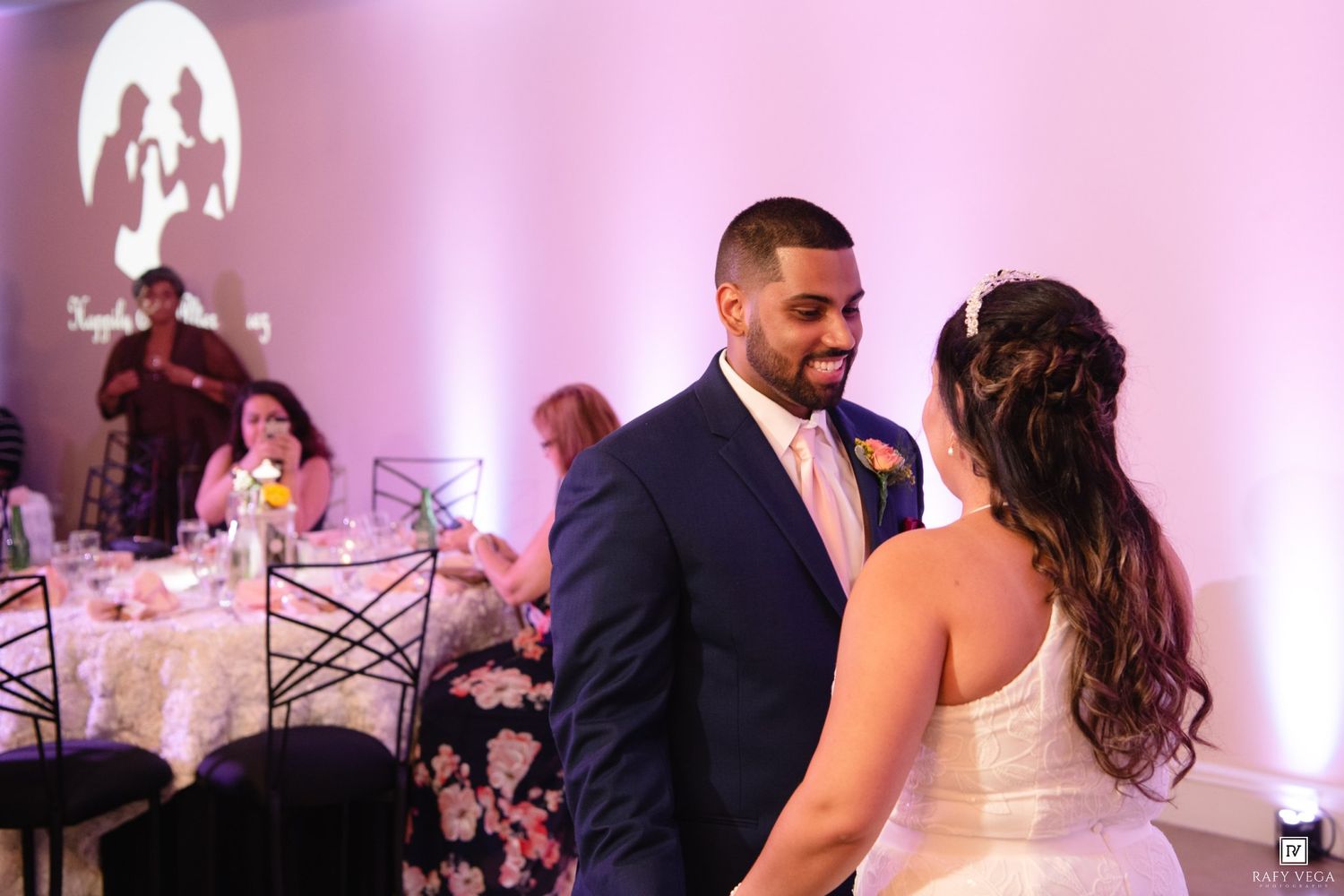 Bride and groom share their first dance at elegant wedding reception with pink uplighting and guests seated in background.