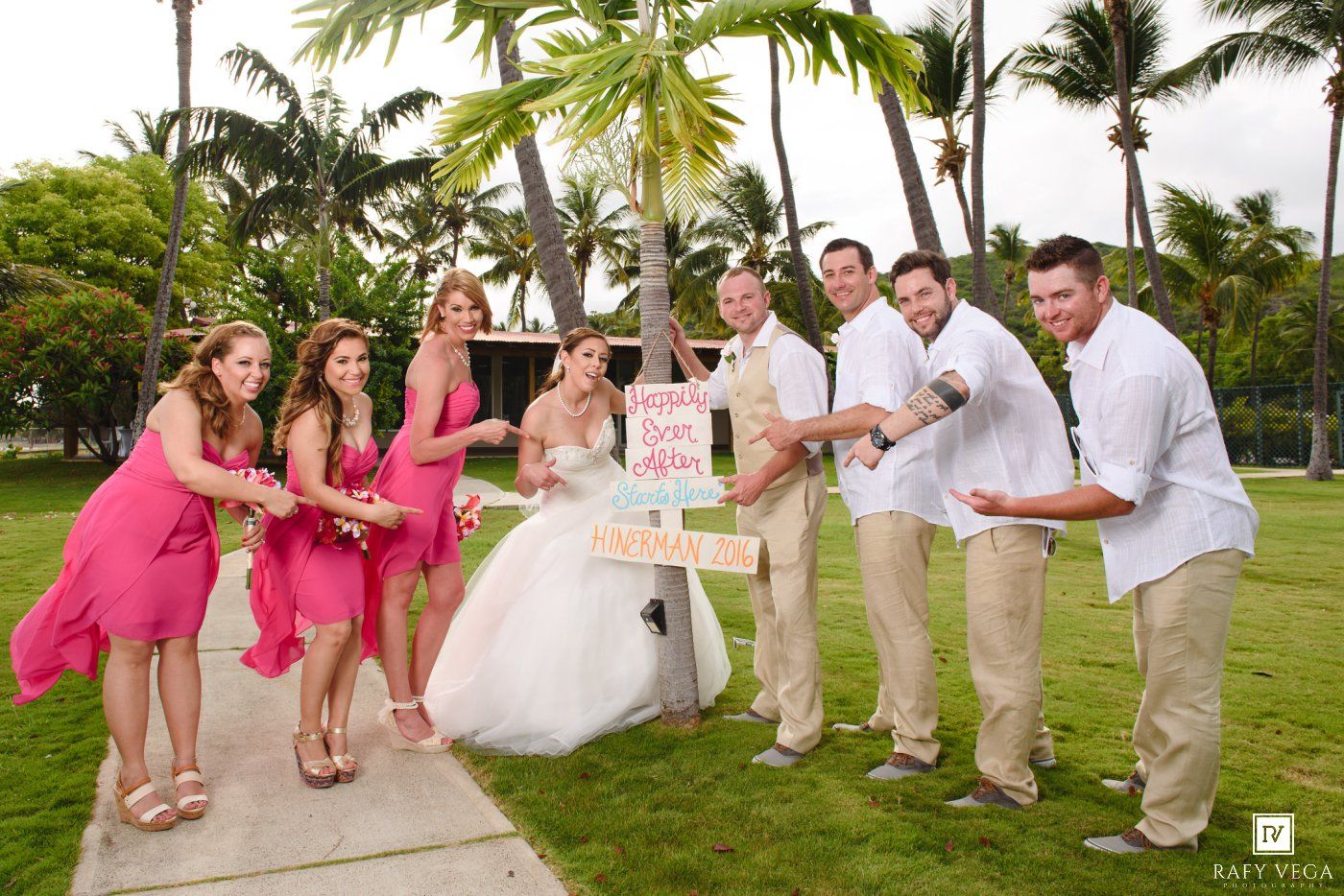 Wedding party in pink bridesmaid dresses and tan suits celebrating outdoors near palm trees at a tropical beach venue.