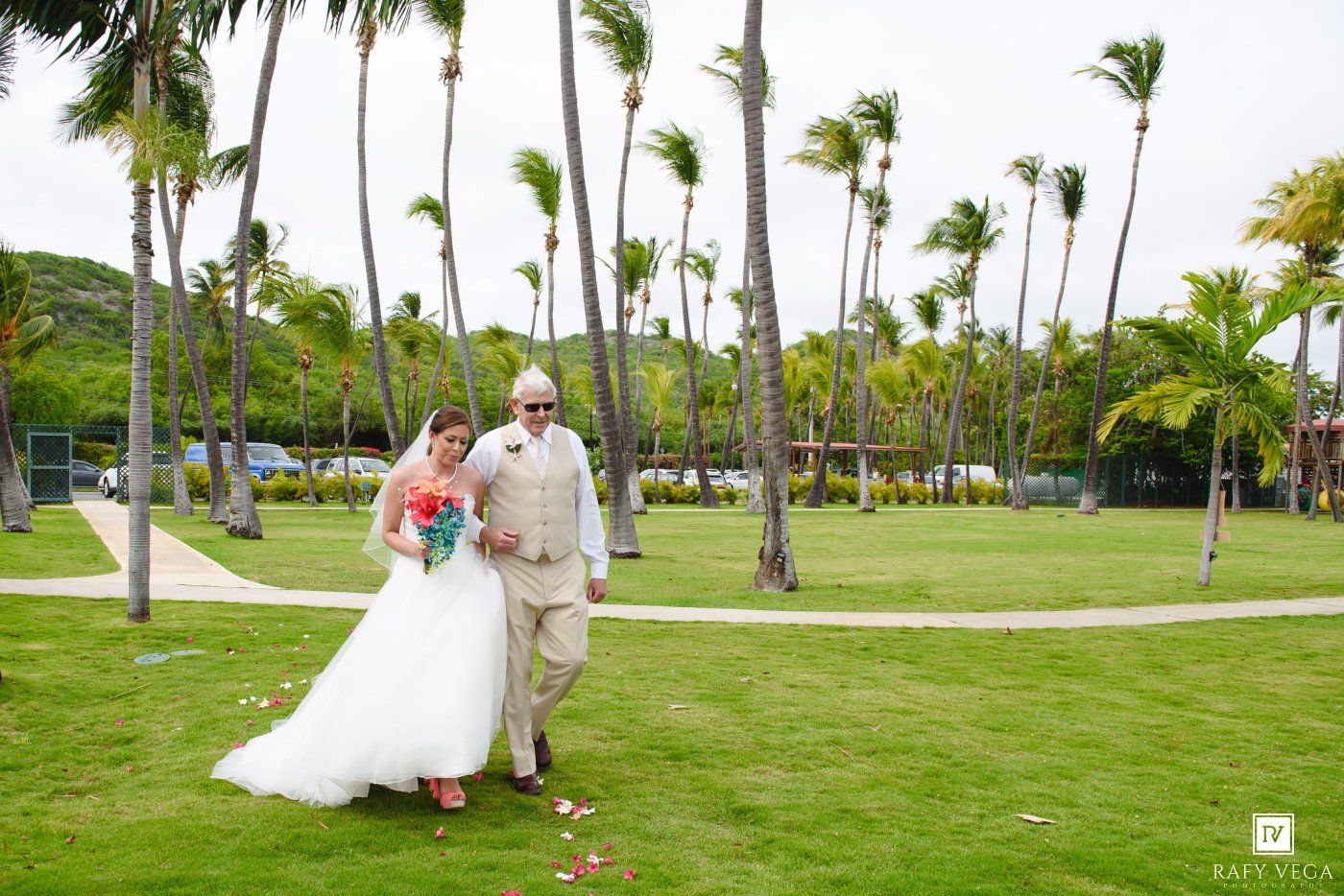 Bride and groom walking together on tropical wedding venue lawn with tall palm trees and lush greenery in background.
