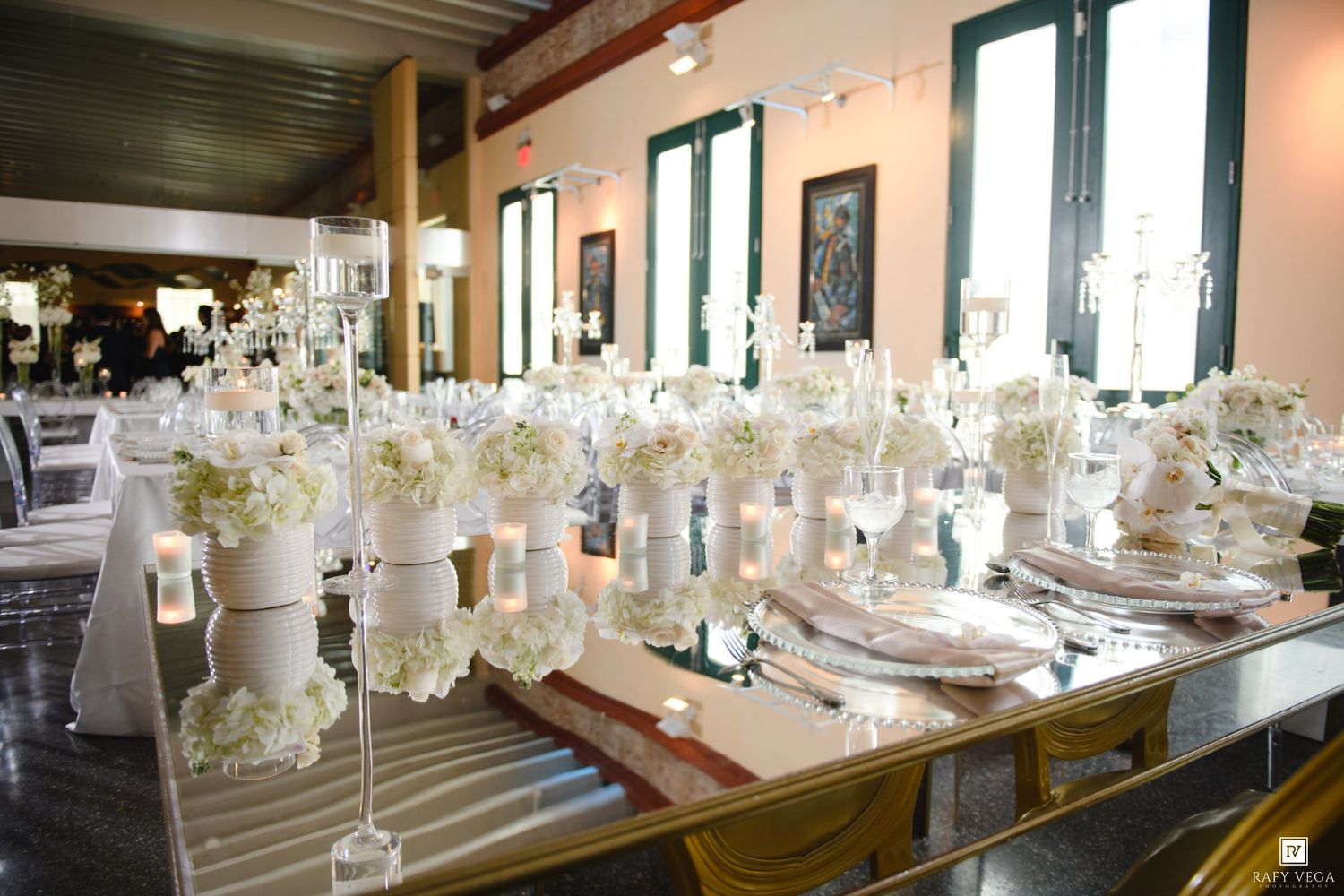 Elegant wedding reception table with white floral centerpieces, candles, and mirror accents in a bright venue.