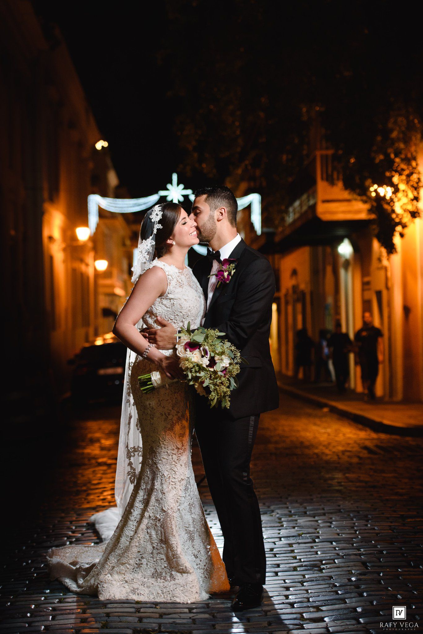 Bride and groom share a romantic kiss on a cobblestone street at night, with glowing lights in the background.