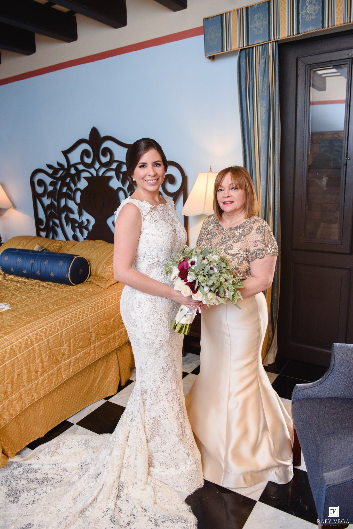 Bride in lace mermaid gown holds bouquet beside woman in champagne dress in elegant hotel room.