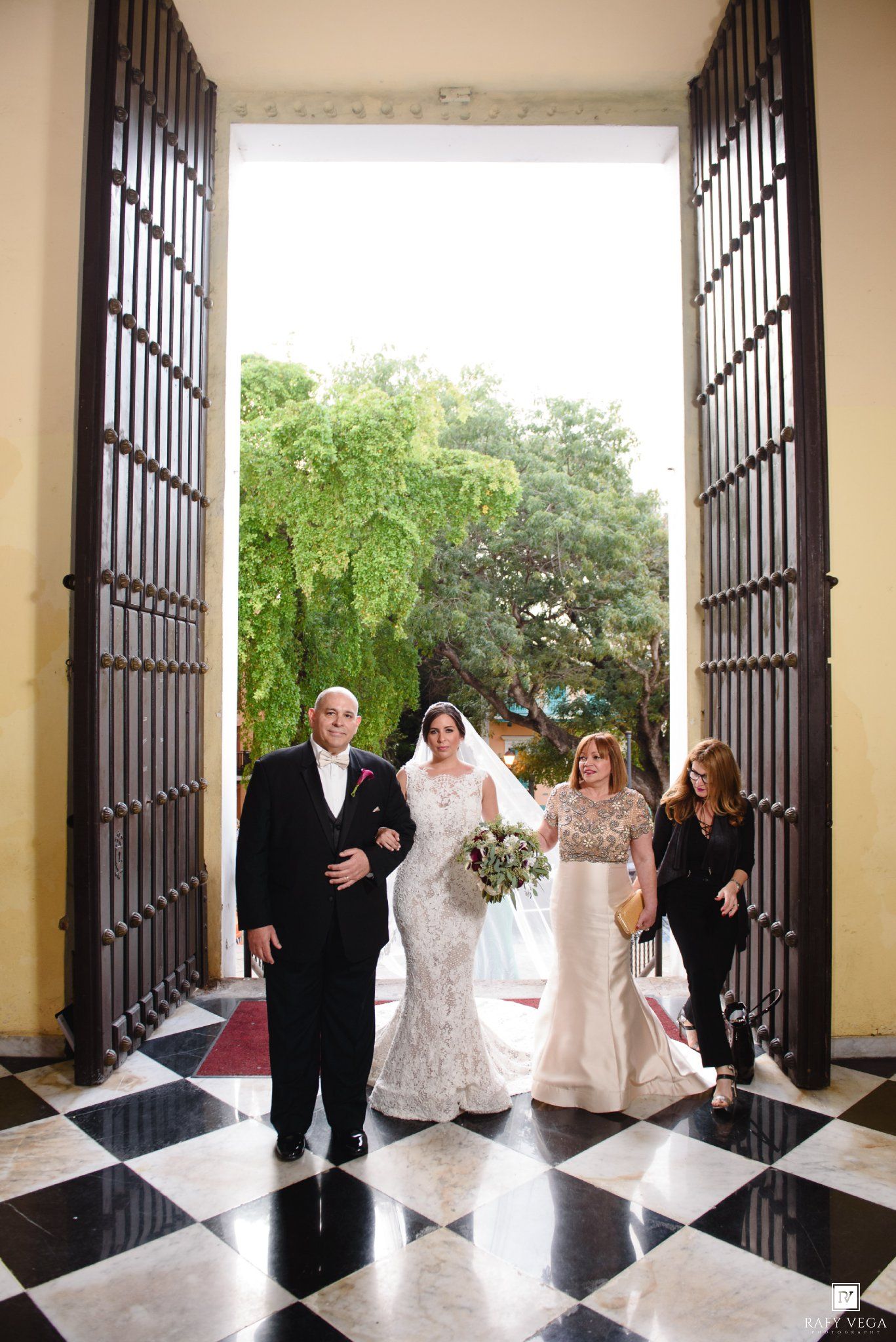 Bride in lace gown entering church with parents through grand wooden doors on wedding day.