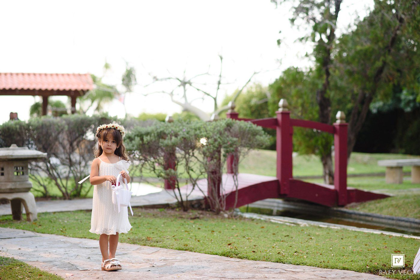 A young flower girl in a white dress walks along a garden path at an outdoor wedding venue.