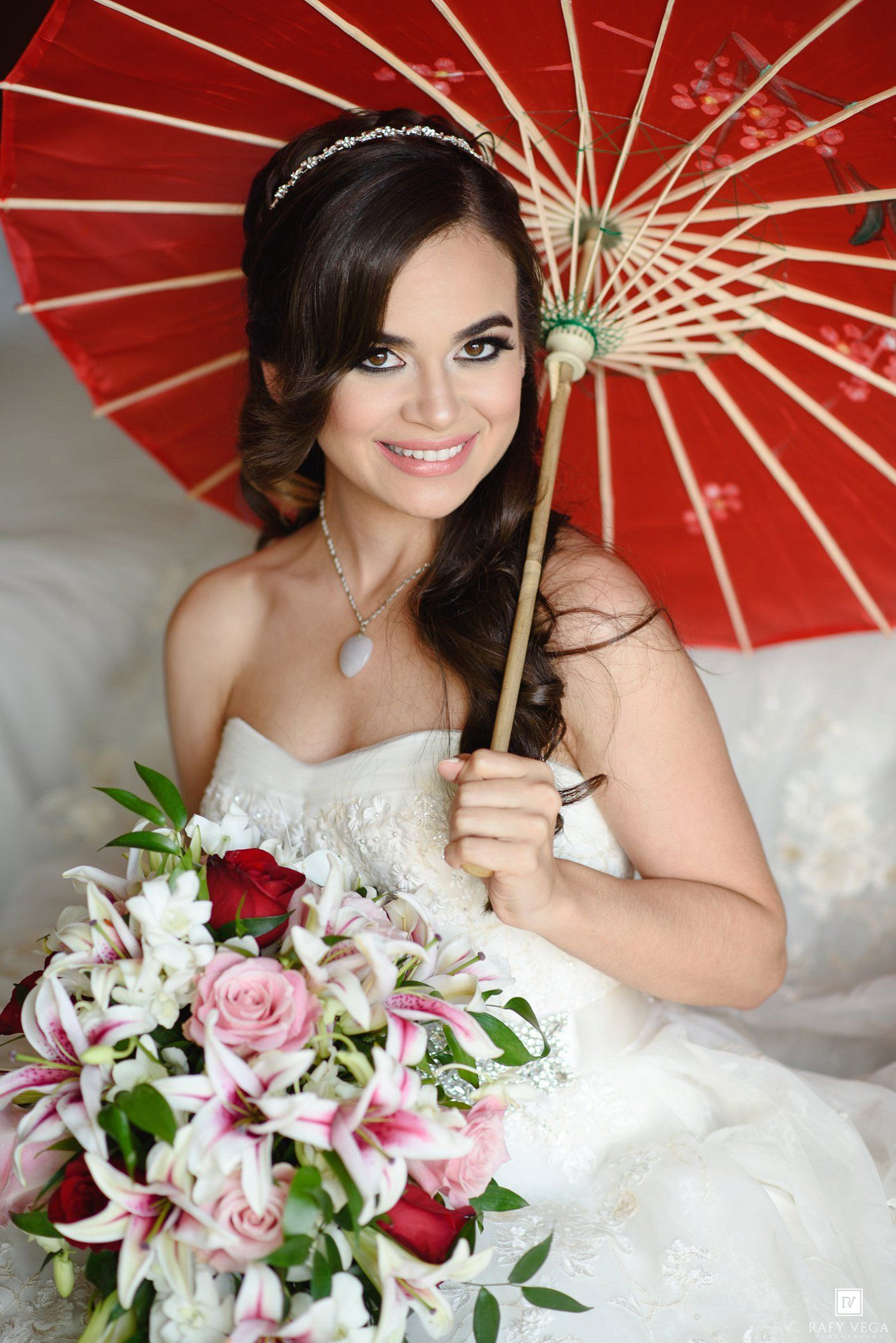 Smiling bride in white gown holding red parasol and pink floral bouquet with lilies and roses.