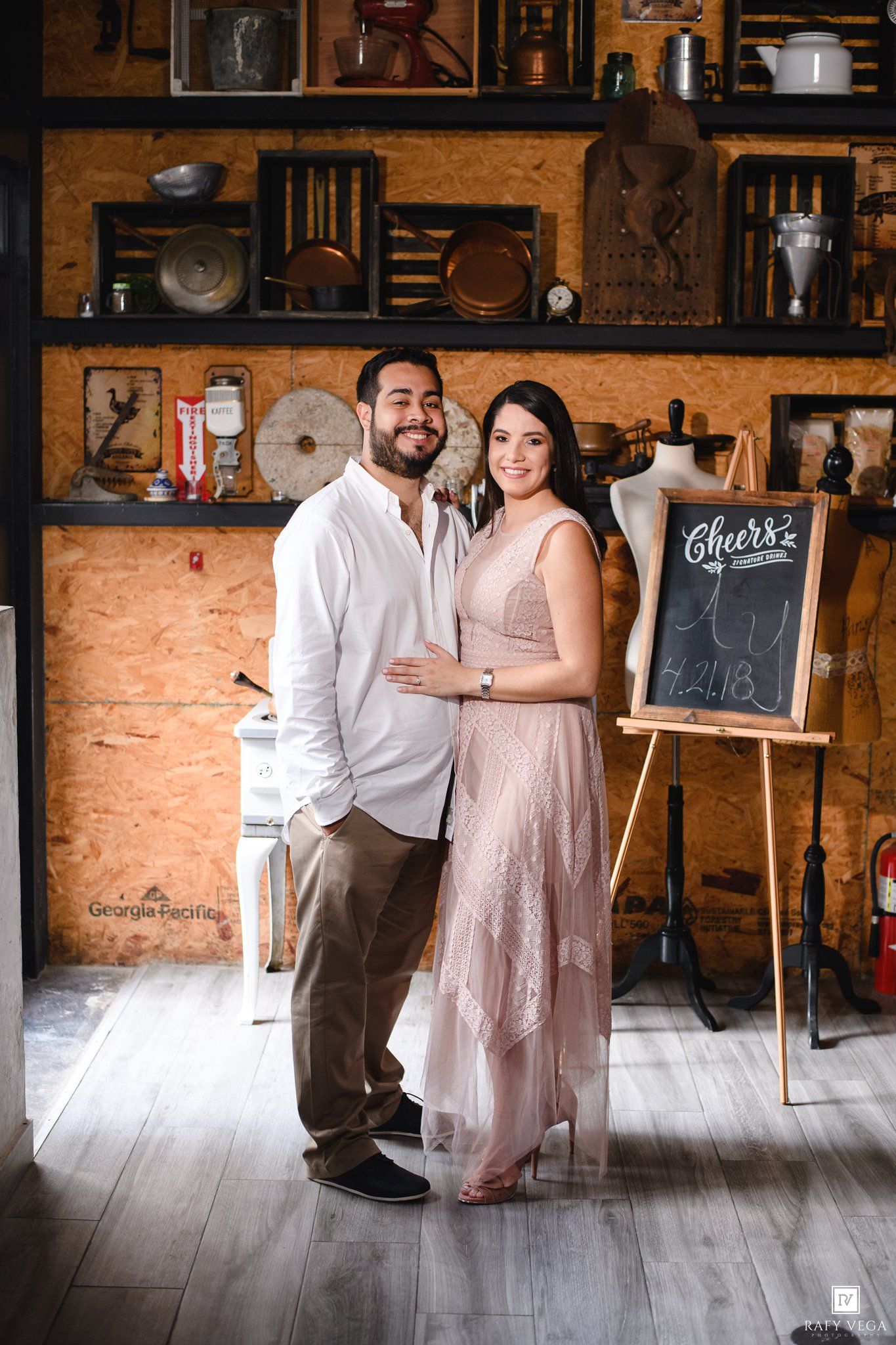 A smiling couple poses together in a rustic studio with vintage decor and a chalkboard sign reading Cheers.