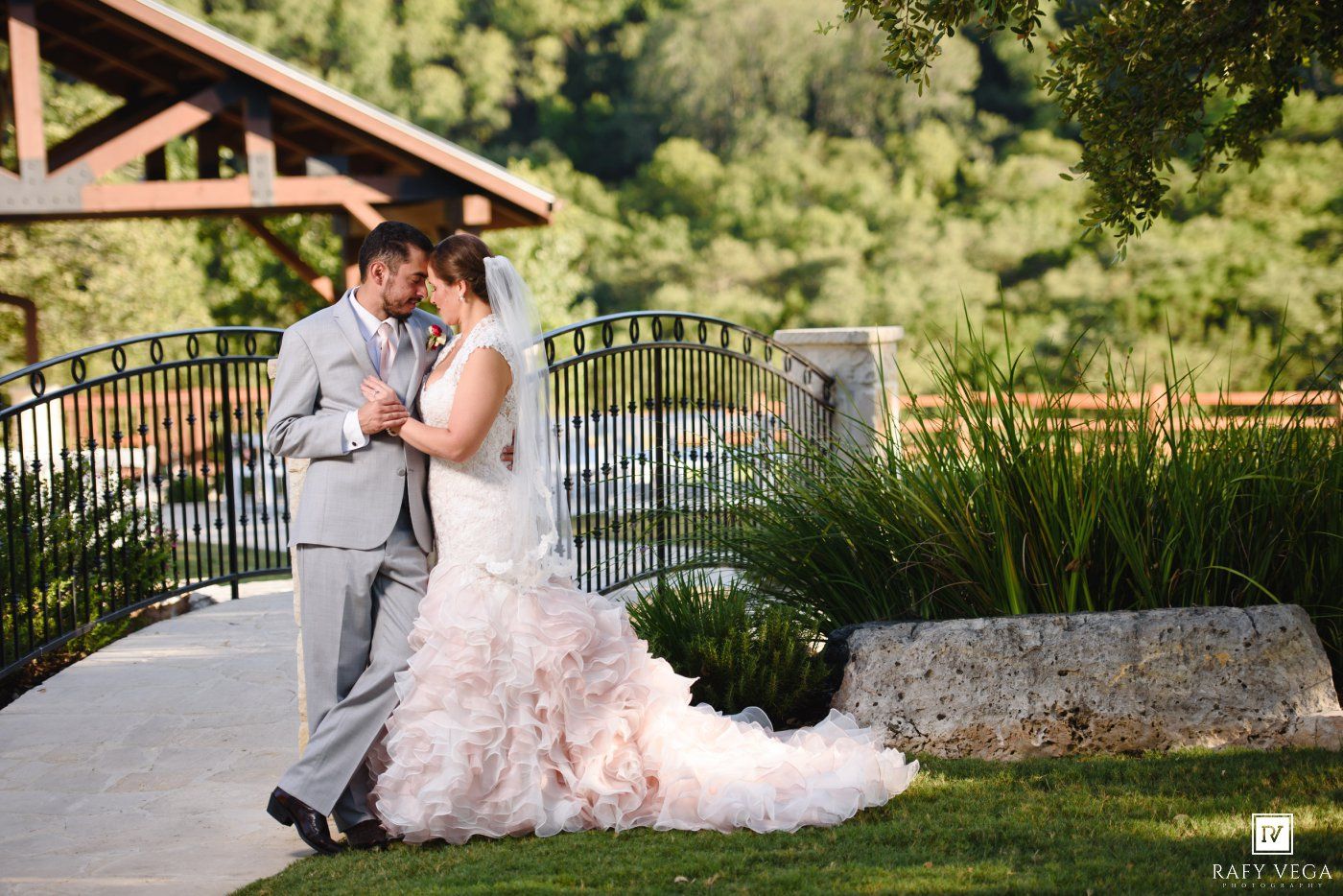 Bride in ruffled blush gown and groom in gray suit share romantic moment on garden bridge surrounded by lush greenery.