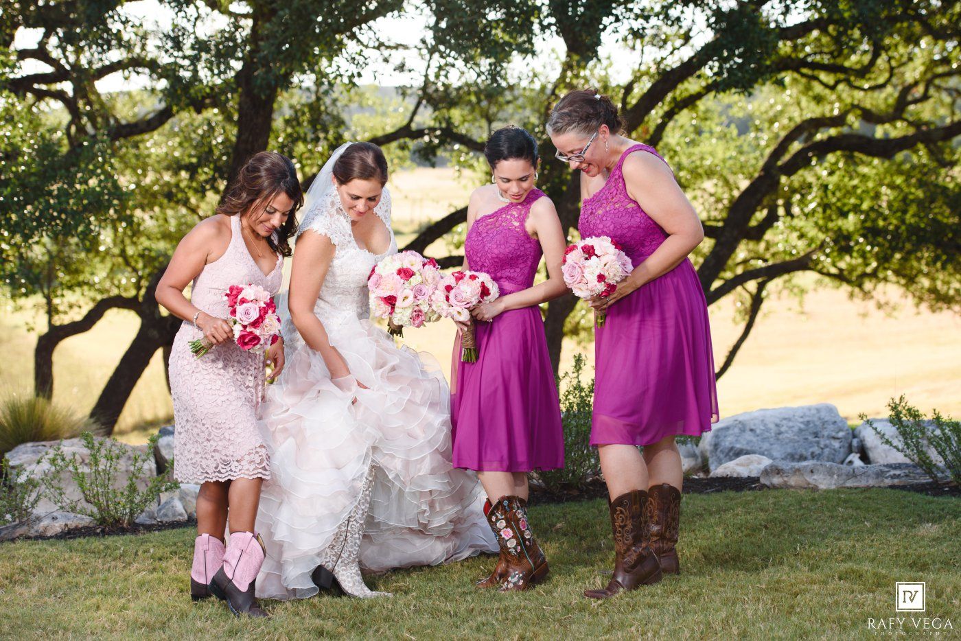 Bridesmaids in purple and pink dresses gather around bride in white gown under oak trees at outdoor wedding.