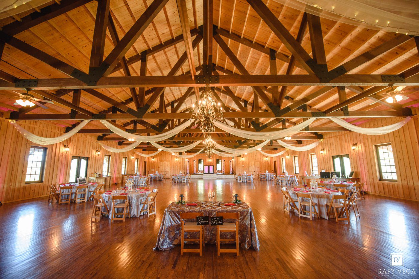 Elegant barn wedding venue with vaulted wood ceilings, draped fabric, chandelier, and round tables set for a reception.