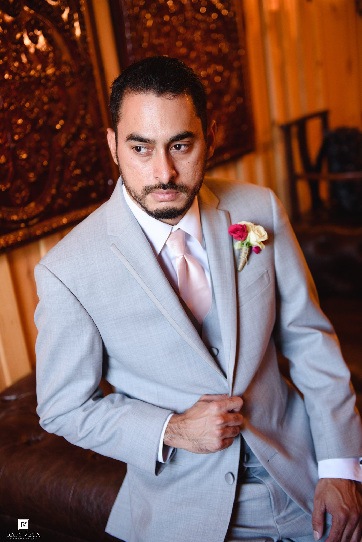 A groom in a light gray suit with a pink tie and floral boutonniere poses confidently indoors.