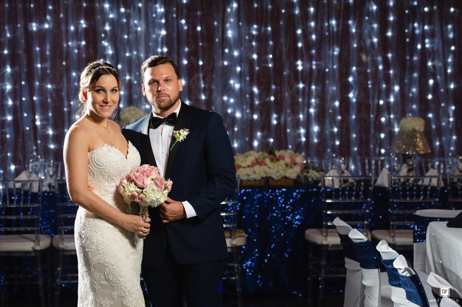 Bride and groom posing together at elegant wedding reception with twinkling fairy light backdrop and blue sequin decor.
