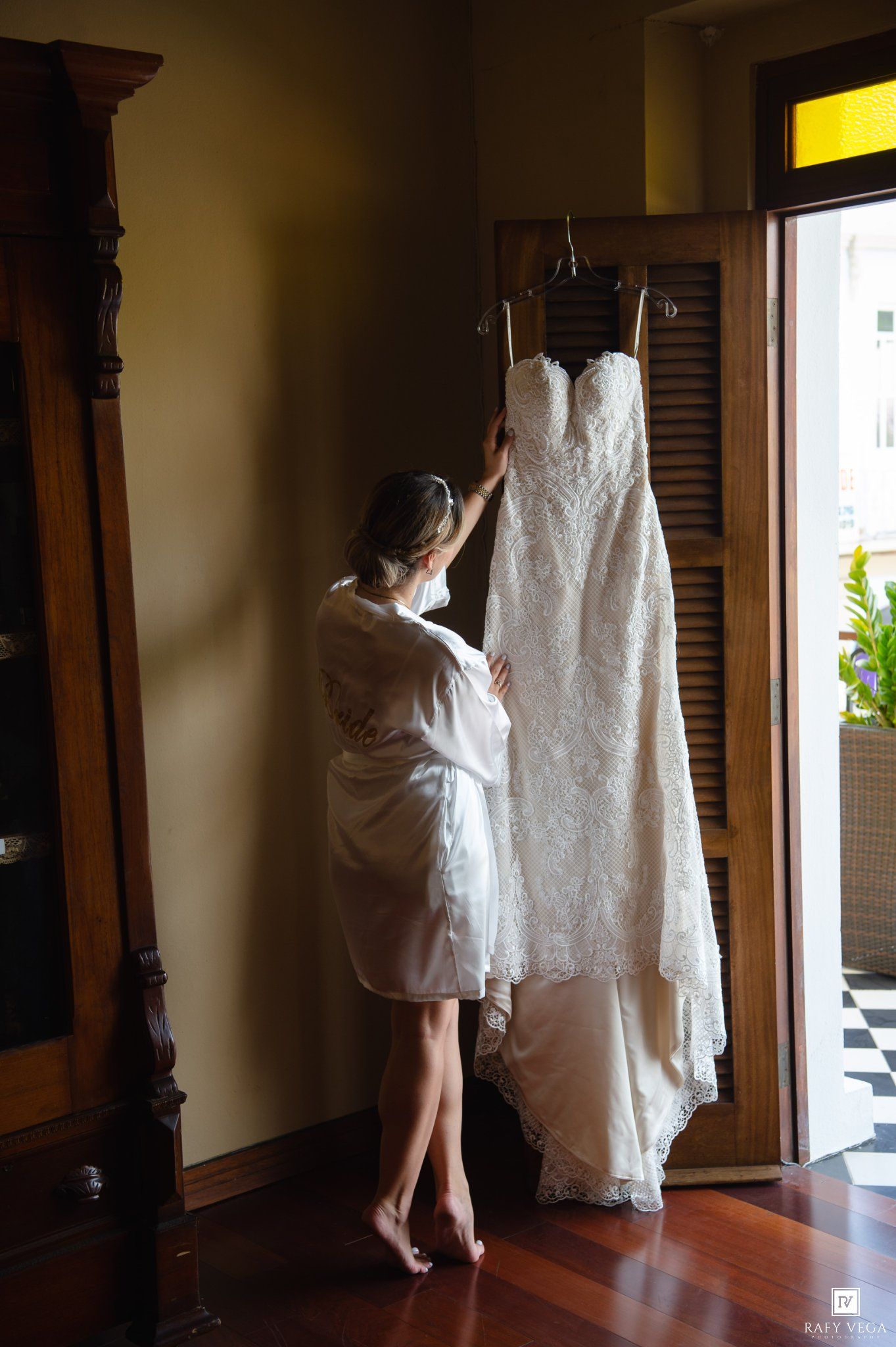Bride in white robe admiring her lace wedding dress hanging by a sunlit doorway on her wedding day.