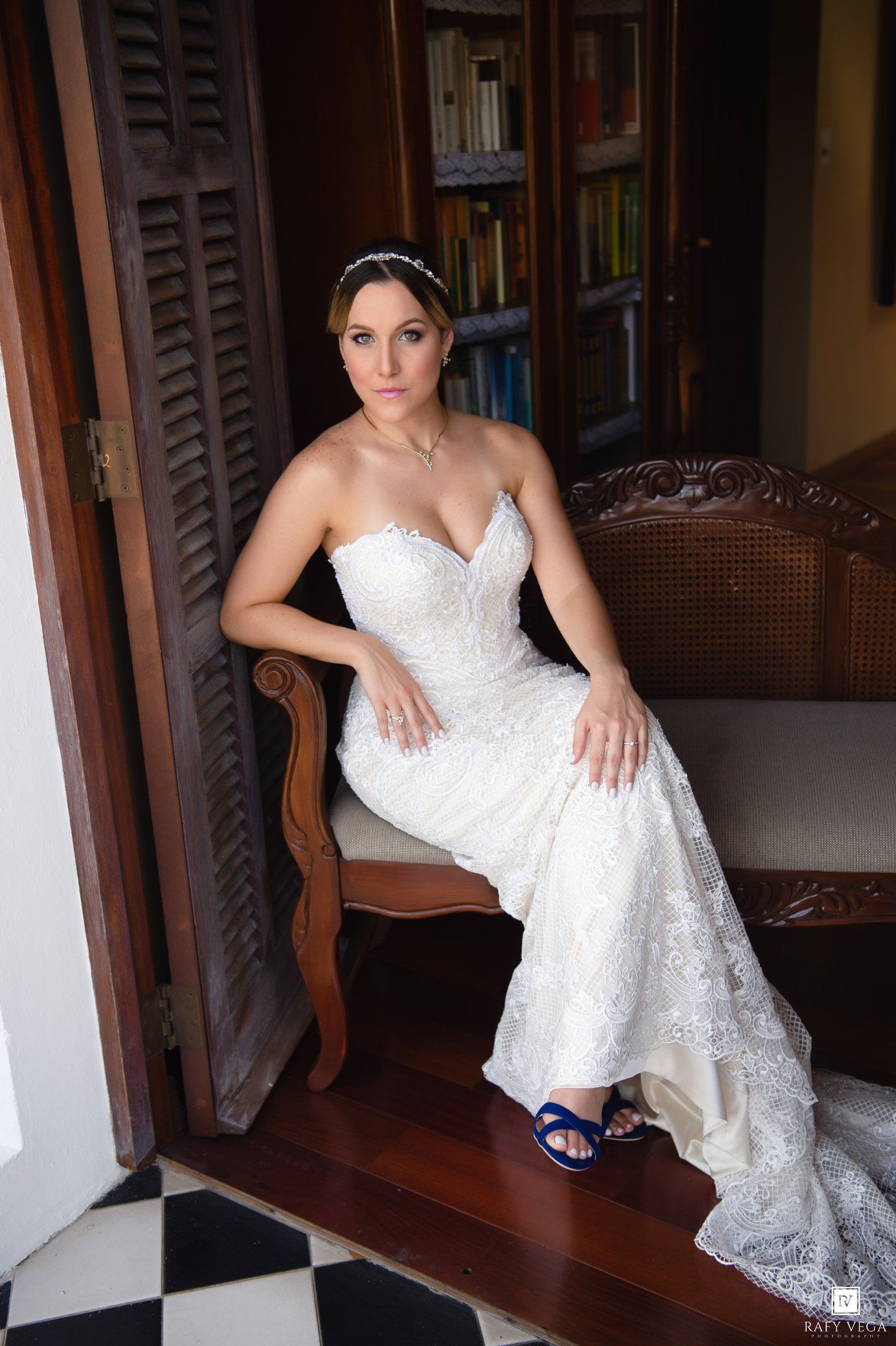 Bride in white lace strapless mermaid gown with blue heels seated on ornate chair in elegant room.