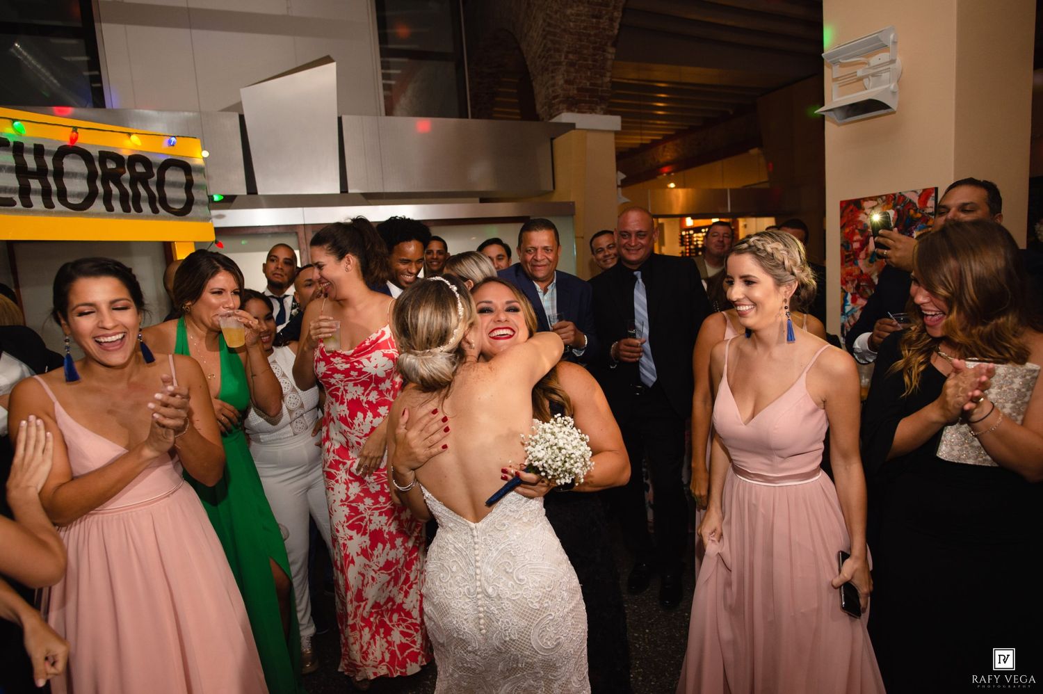 Bride in white gown hugging guest at wedding reception surrounded by bridesmaids in pink dresses.