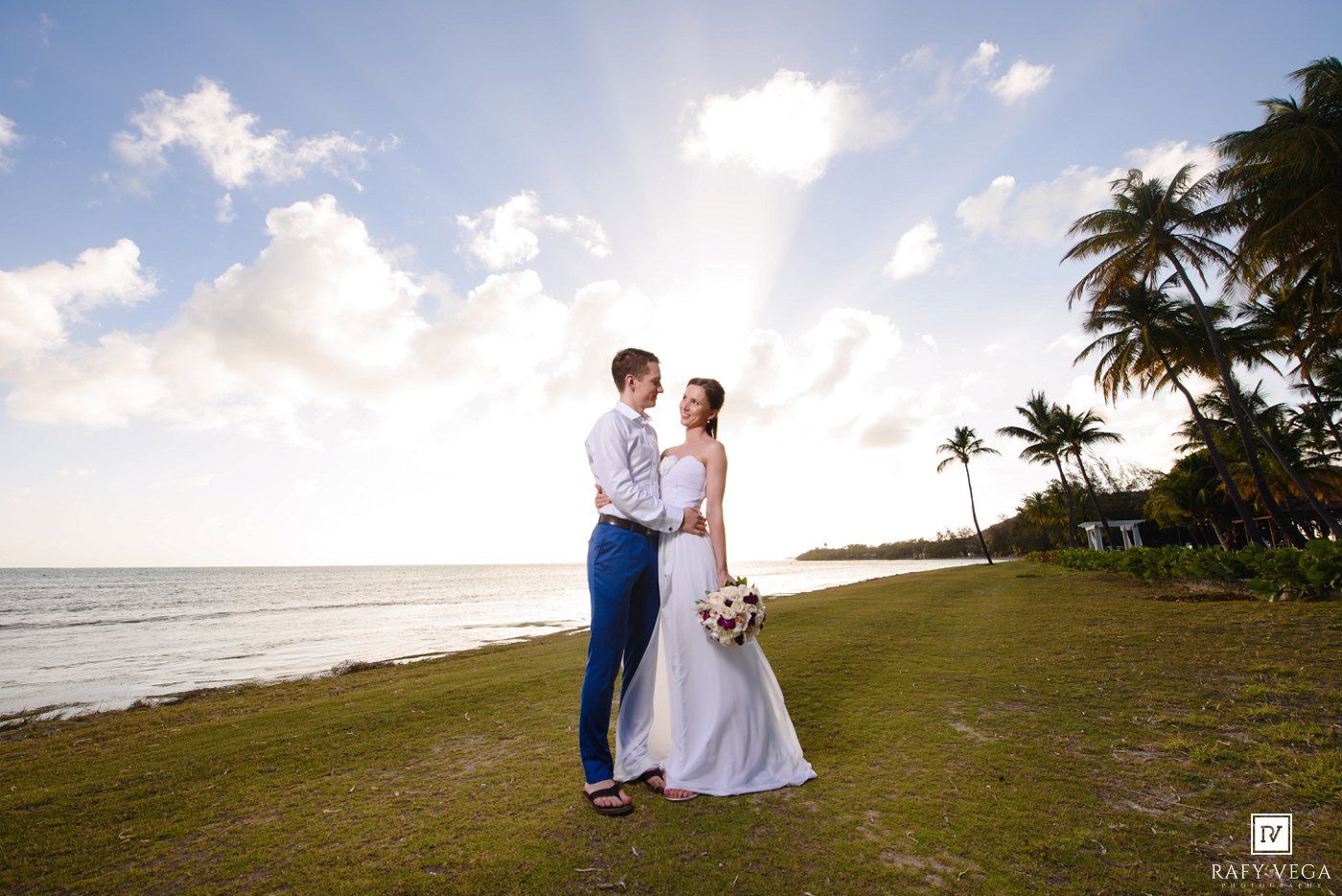 Bride and groom share a romantic moment on a tropical beach at sunset with palm trees and ocean views.