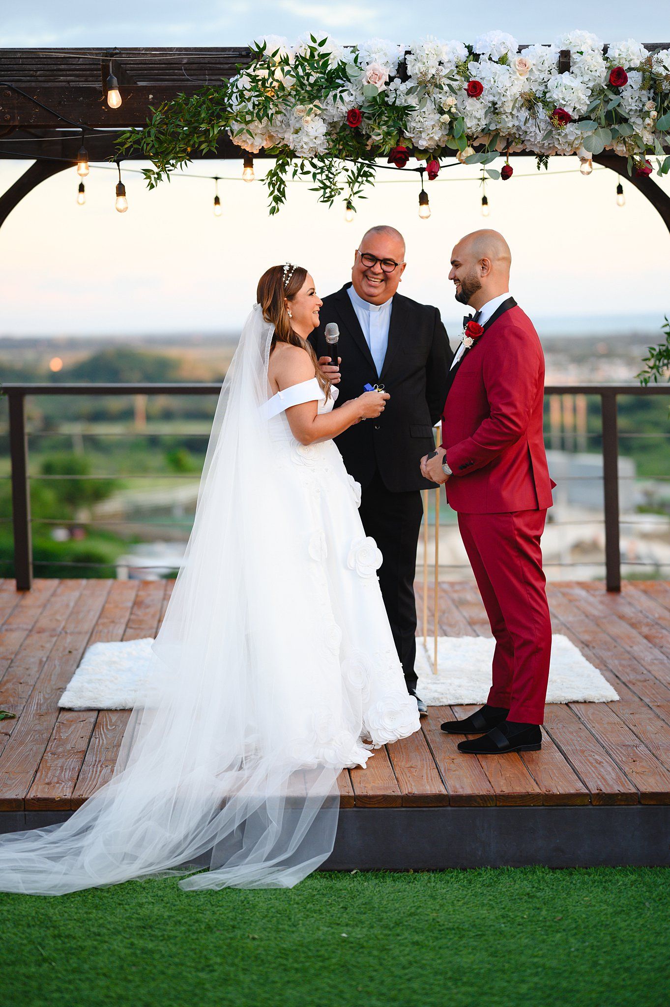 Bride in white gown and groom in red suit exchange vows outdoors under floral arch at sunset wedding ceremony.