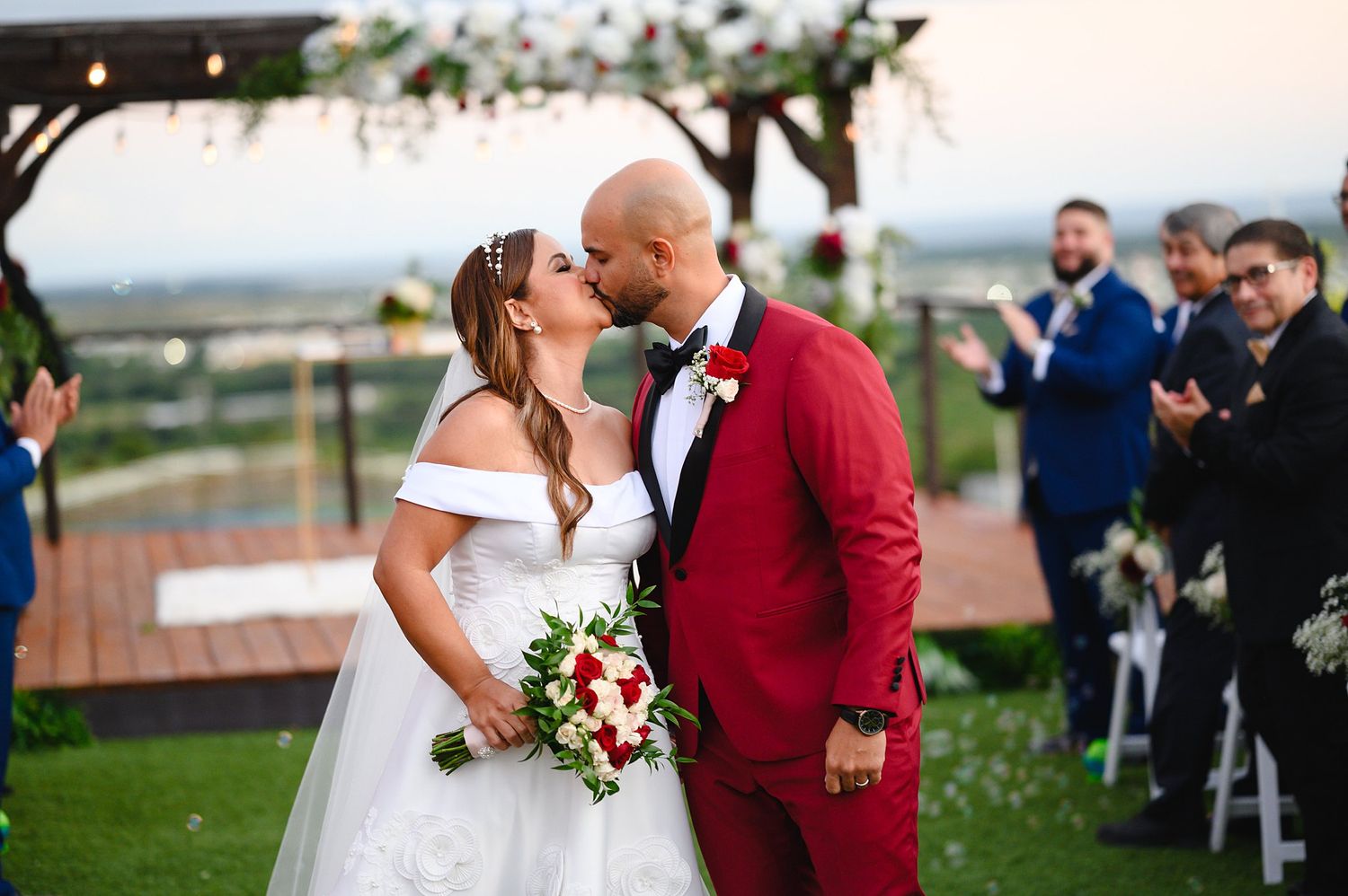 Bride and groom share a kiss at outdoor wedding ceremony, groom in red suit holding bouquet of red and white flowers.