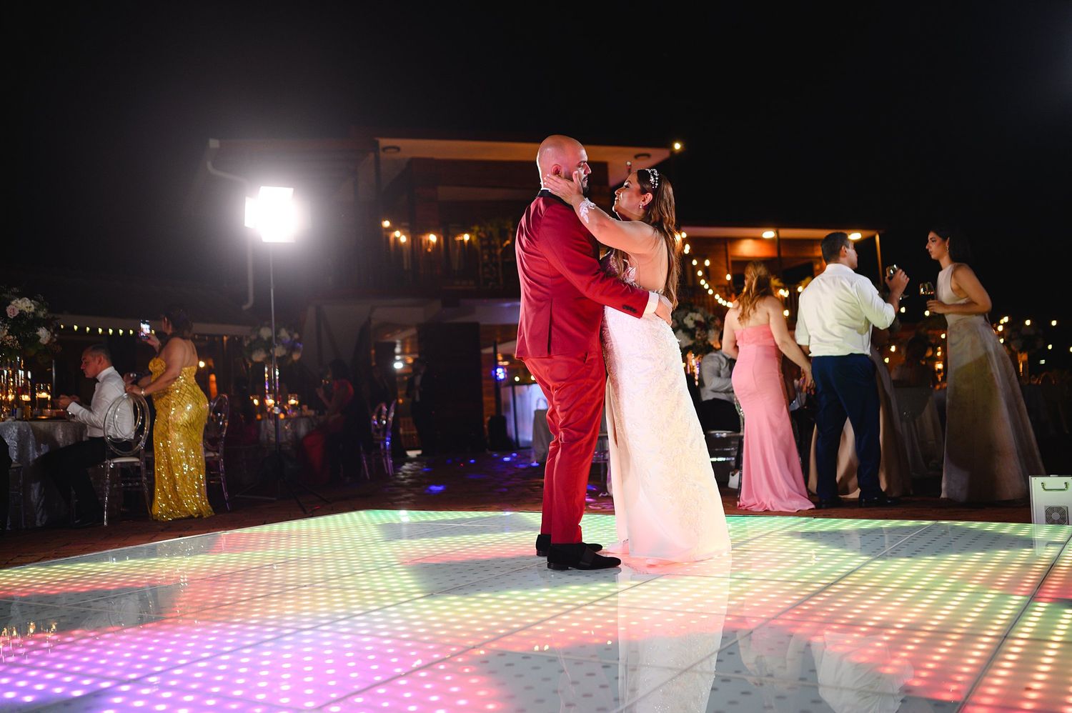 Couple shares first dance on colorful LED dance floor at outdoor wedding reception at night.
