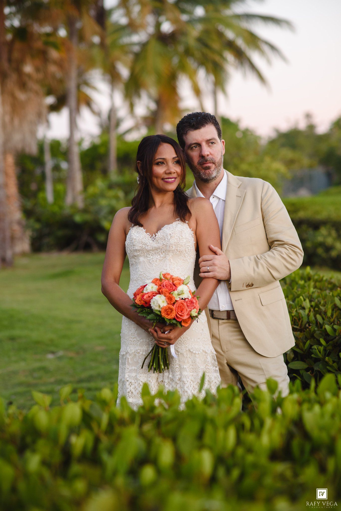 Bride and groom pose outdoors at tropical wedding, bride holds orange rose bouquet, groom wears tan suit.