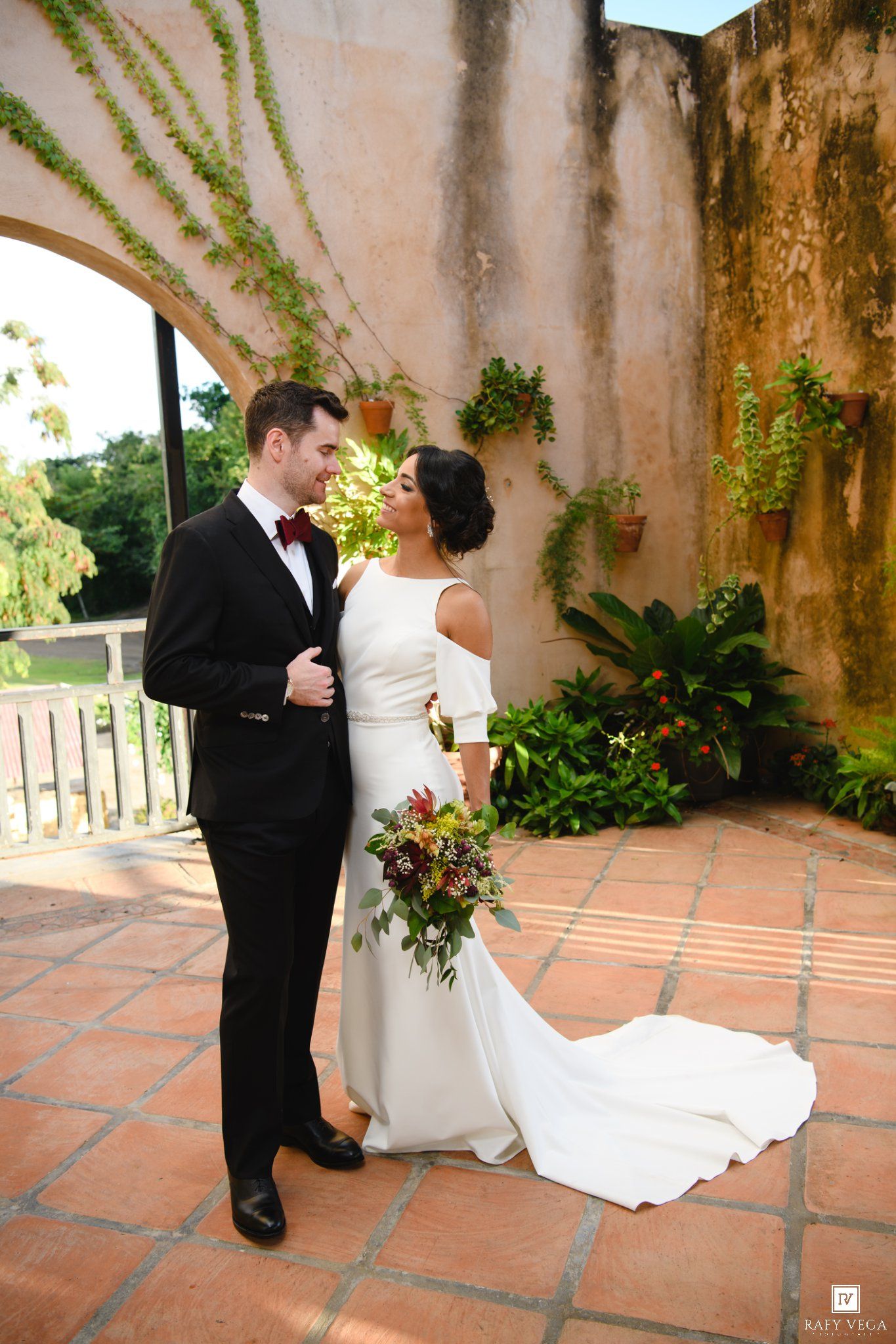 Bride and groom share a romantic moment in a lush courtyard, she holds a burgundy bouquet in a white gown.